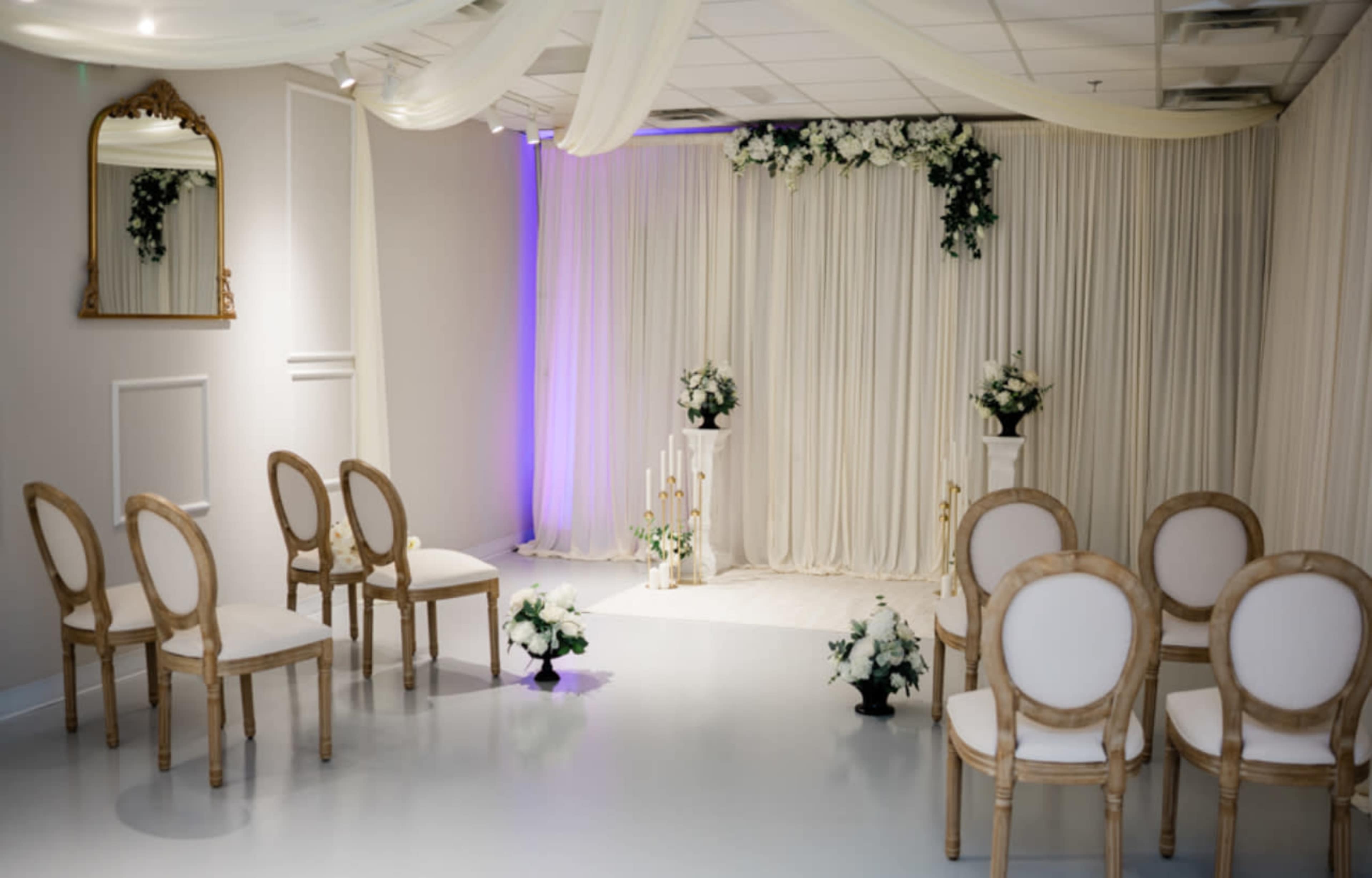 The image shows a decorated event space featuring white chairs arranged in rows, a backdrop of draped fabric with floral arrangements, and candles in a central display.