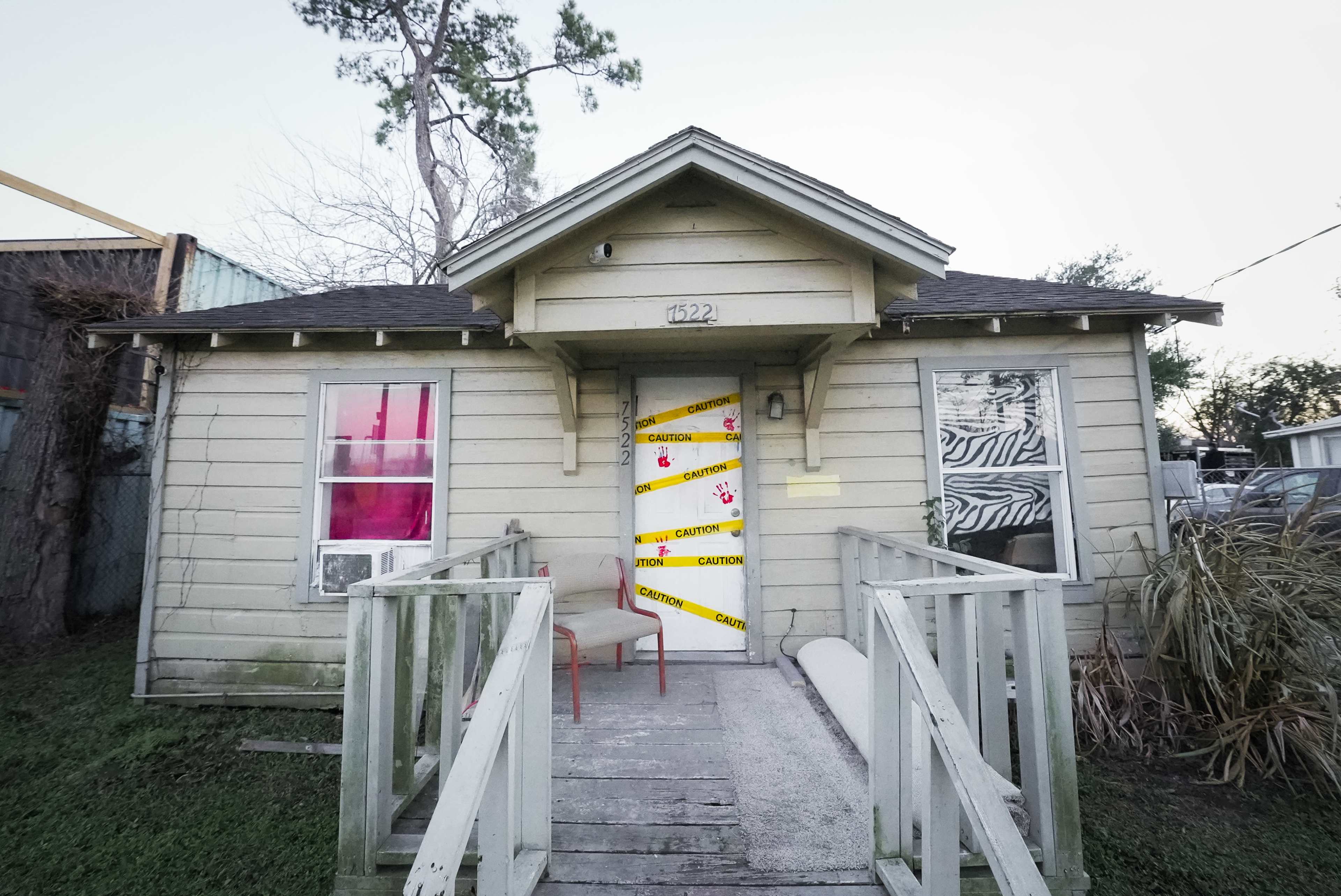 The image displays a small, weathered house with caution tape across the front door and a single chair placed on the porch.