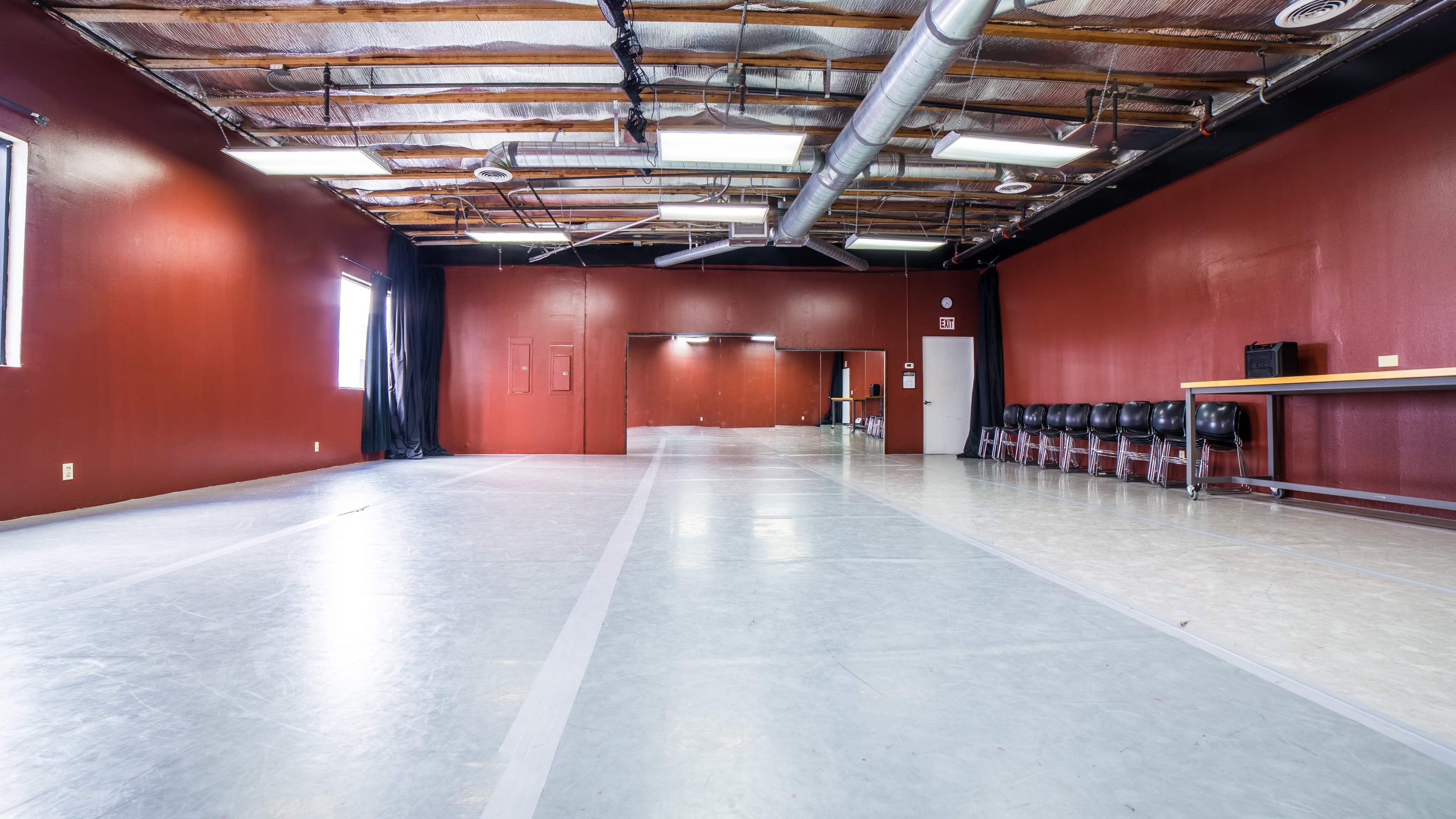 An empty studio space features a polished floor and red walls, with a row of black chairs along one side.