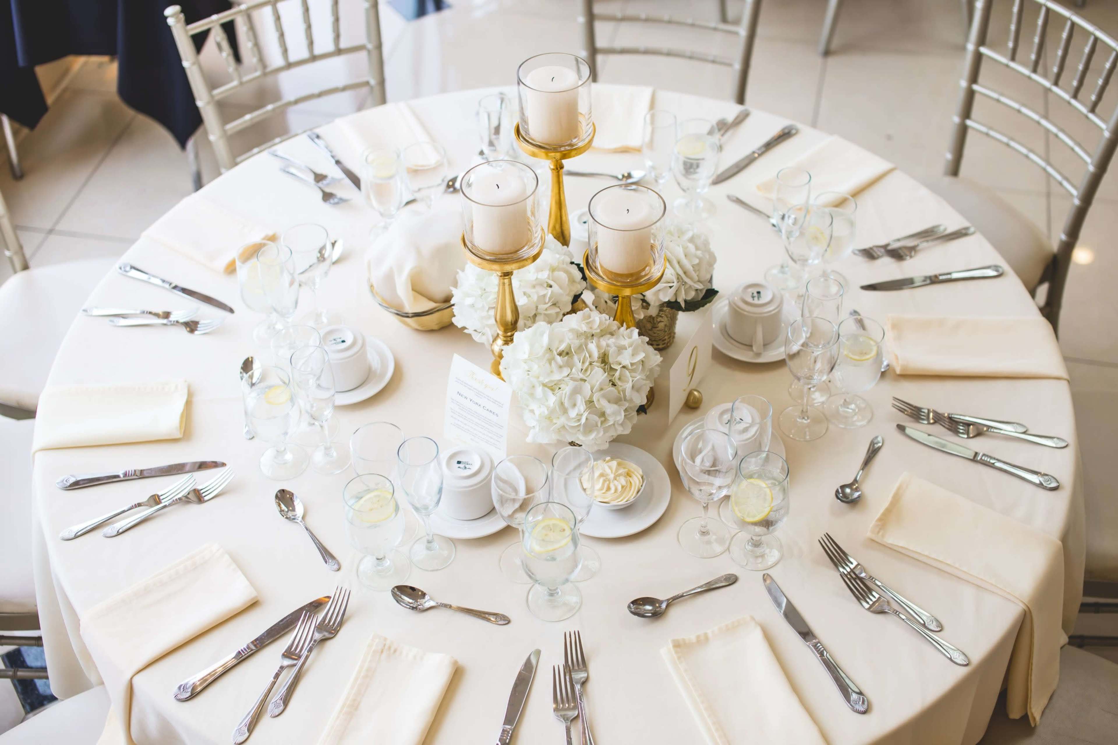 A elegantly set dining table with white linens features floral centerpieces, candles, and arranged cutlery.
