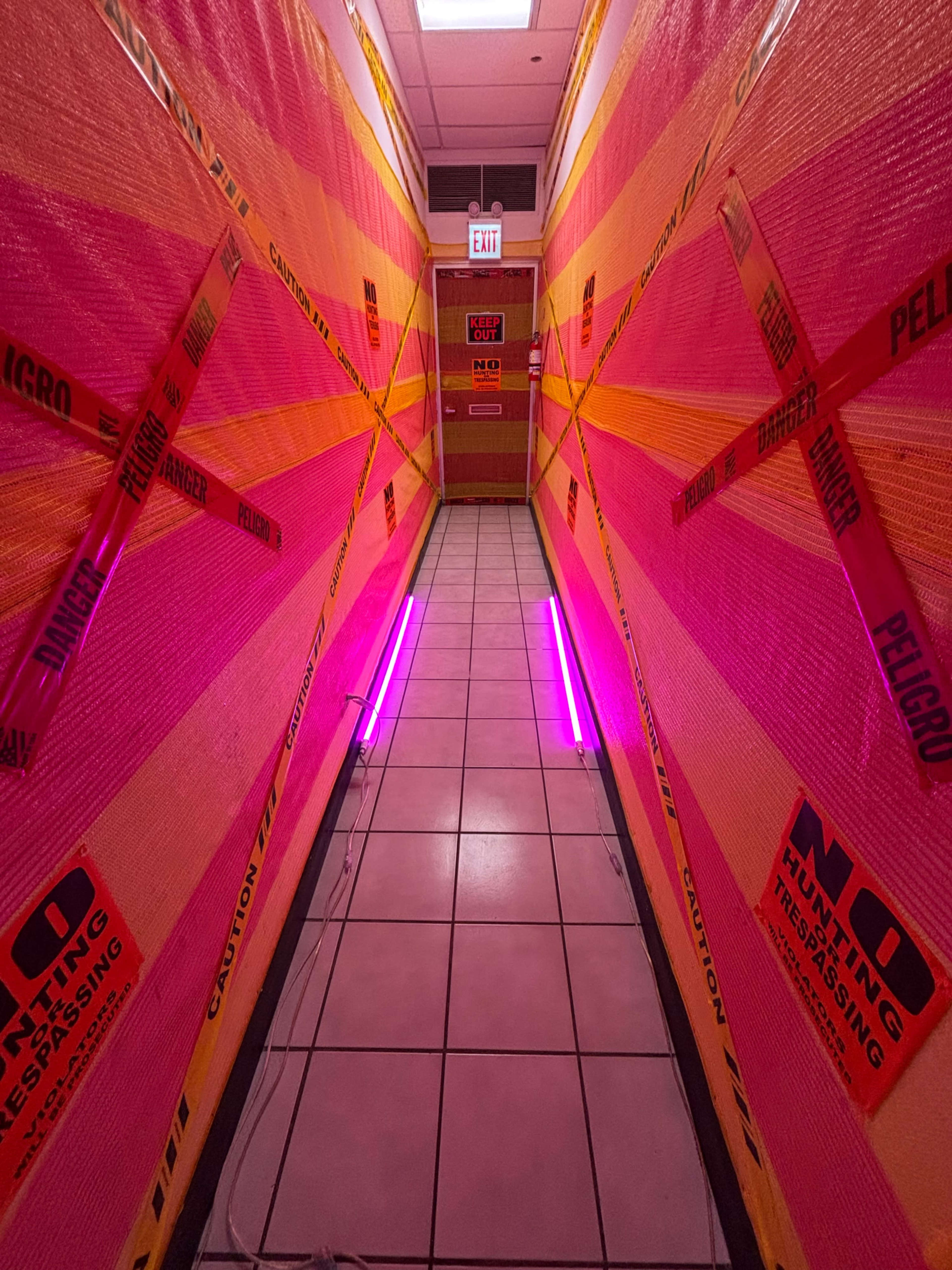 The image shows a corridor lined with bright pink and orange caution tape, with fluorescent lights illuminating the tiled floor.