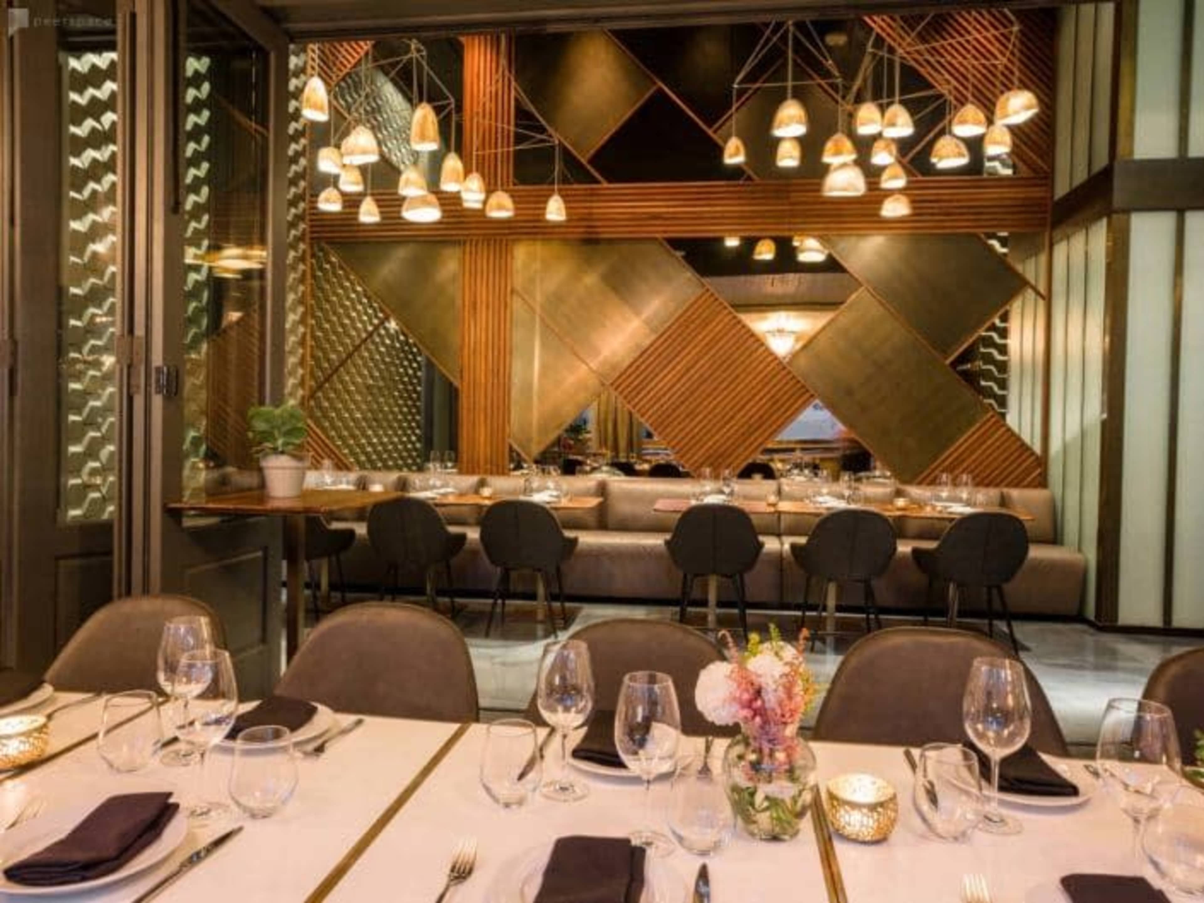 A modern restaurant interior with a long dining table set with glassware and napkins, surrounded by decorative light fixtures and geometric wall designs.