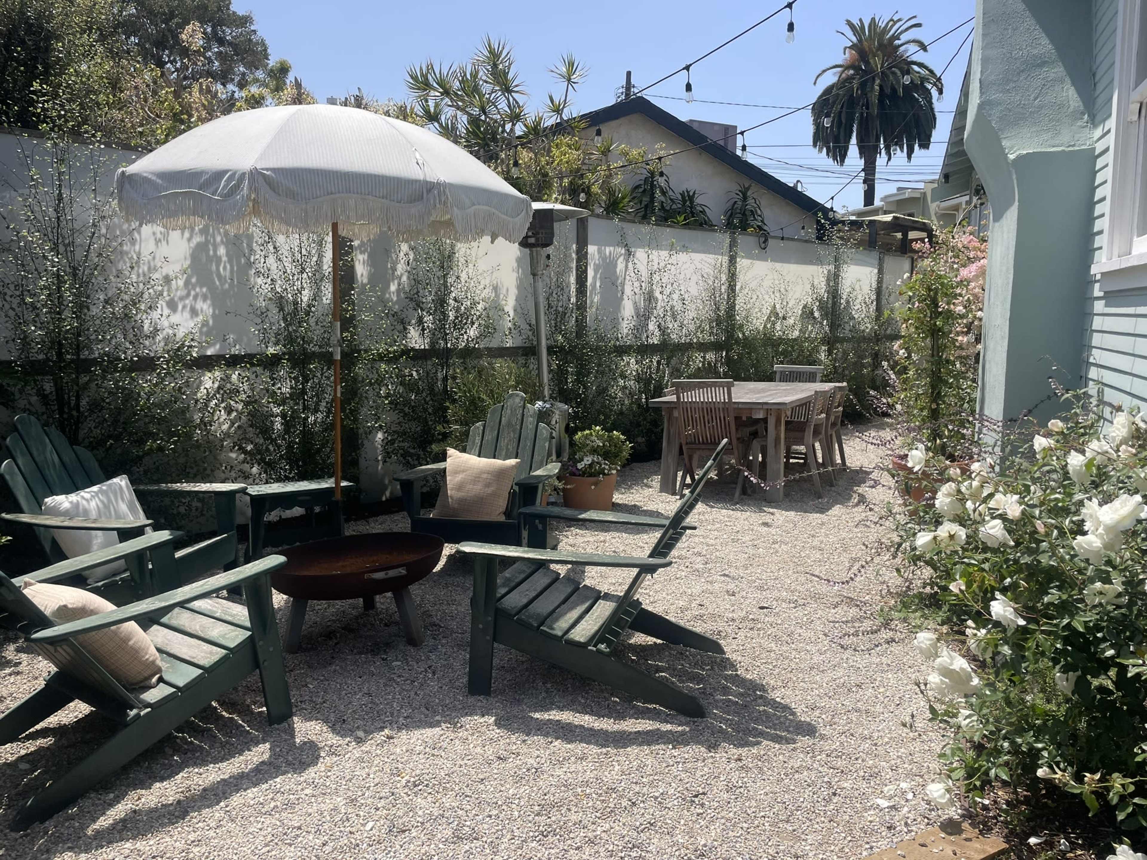 The image shows a landscaped outdoor area with green patio chairs, a table, an umbrella, and a gravel surface surrounded by plants and a white wall.