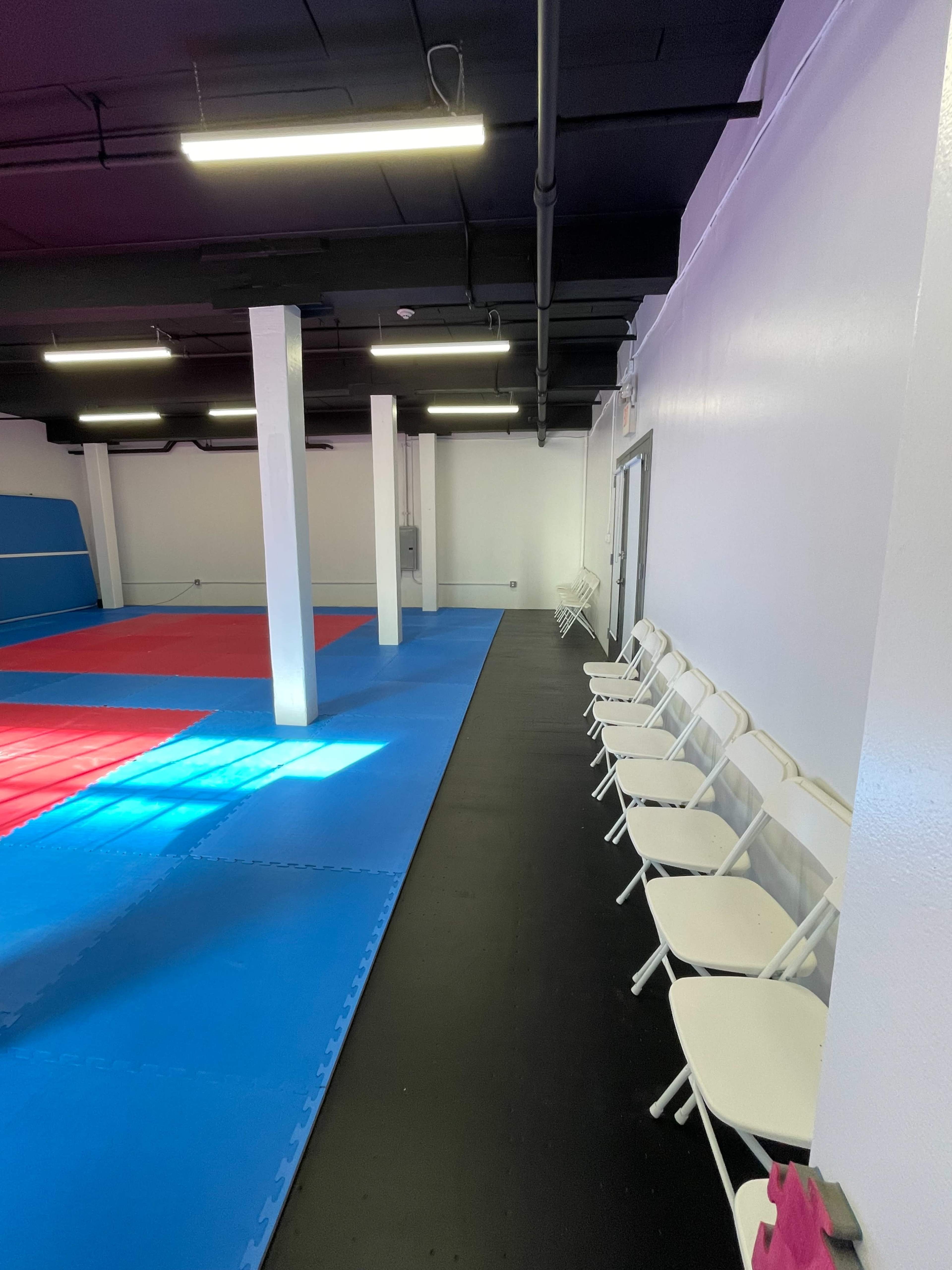 An empty martial arts training facility with a blue and red mat area and a row of folding chairs along one side.
