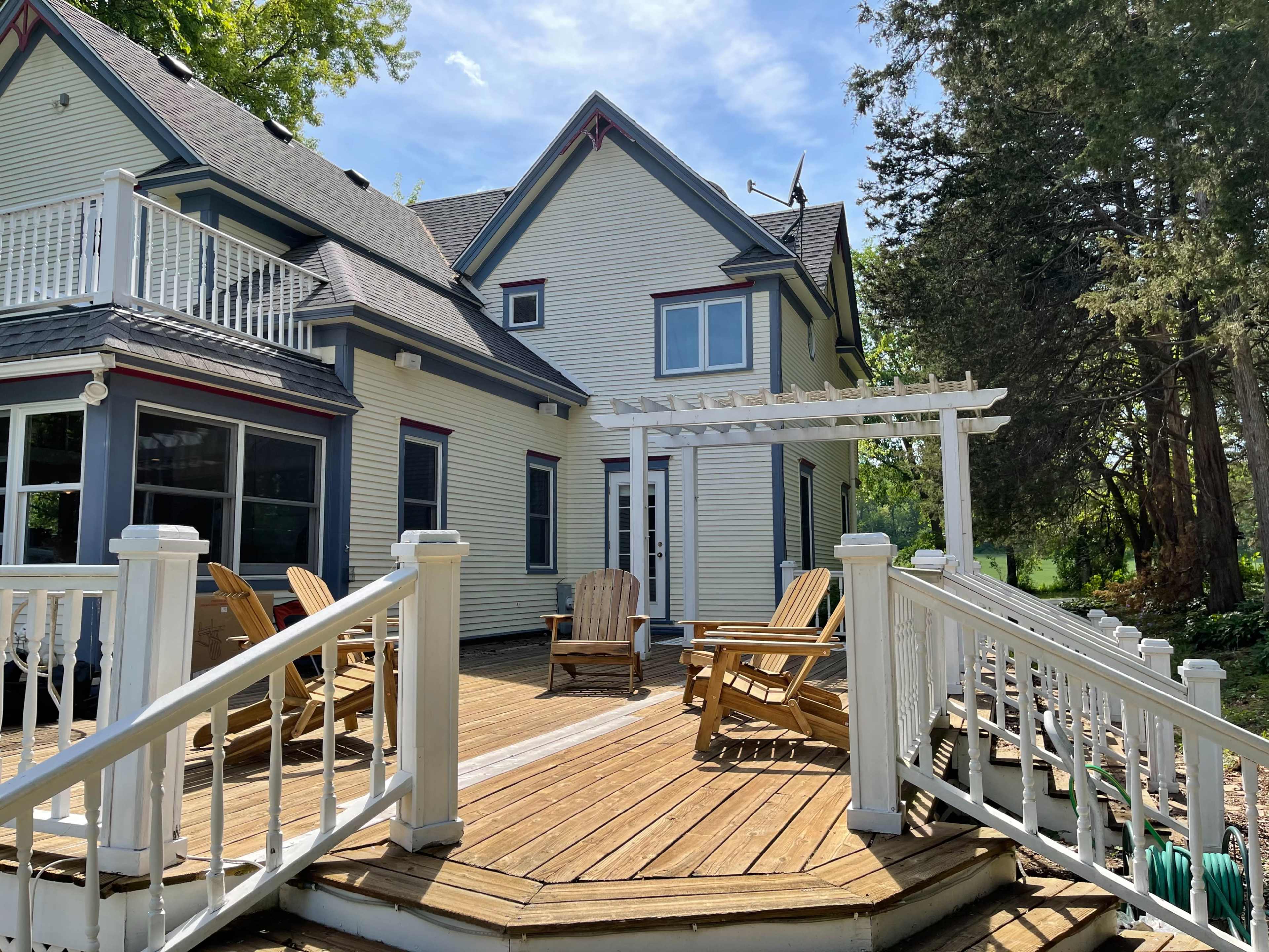 A large house with a deck featuring wooden chairs and a pergola, surrounded by greenery under a bright sky.