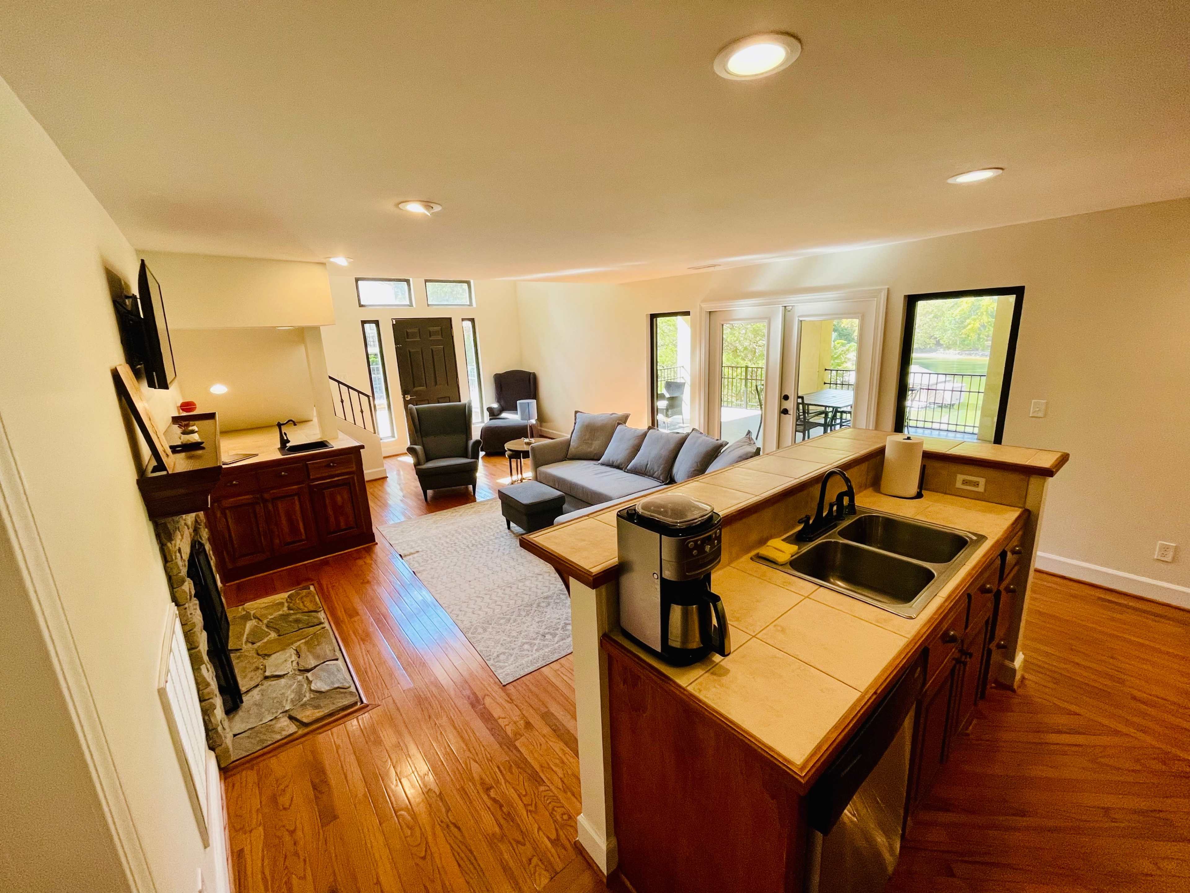 The image shows a spacious living area with a kitchen bar, featuring a gray sofa set, a stone fireplace, and large windows overlooking an outdoor patio.