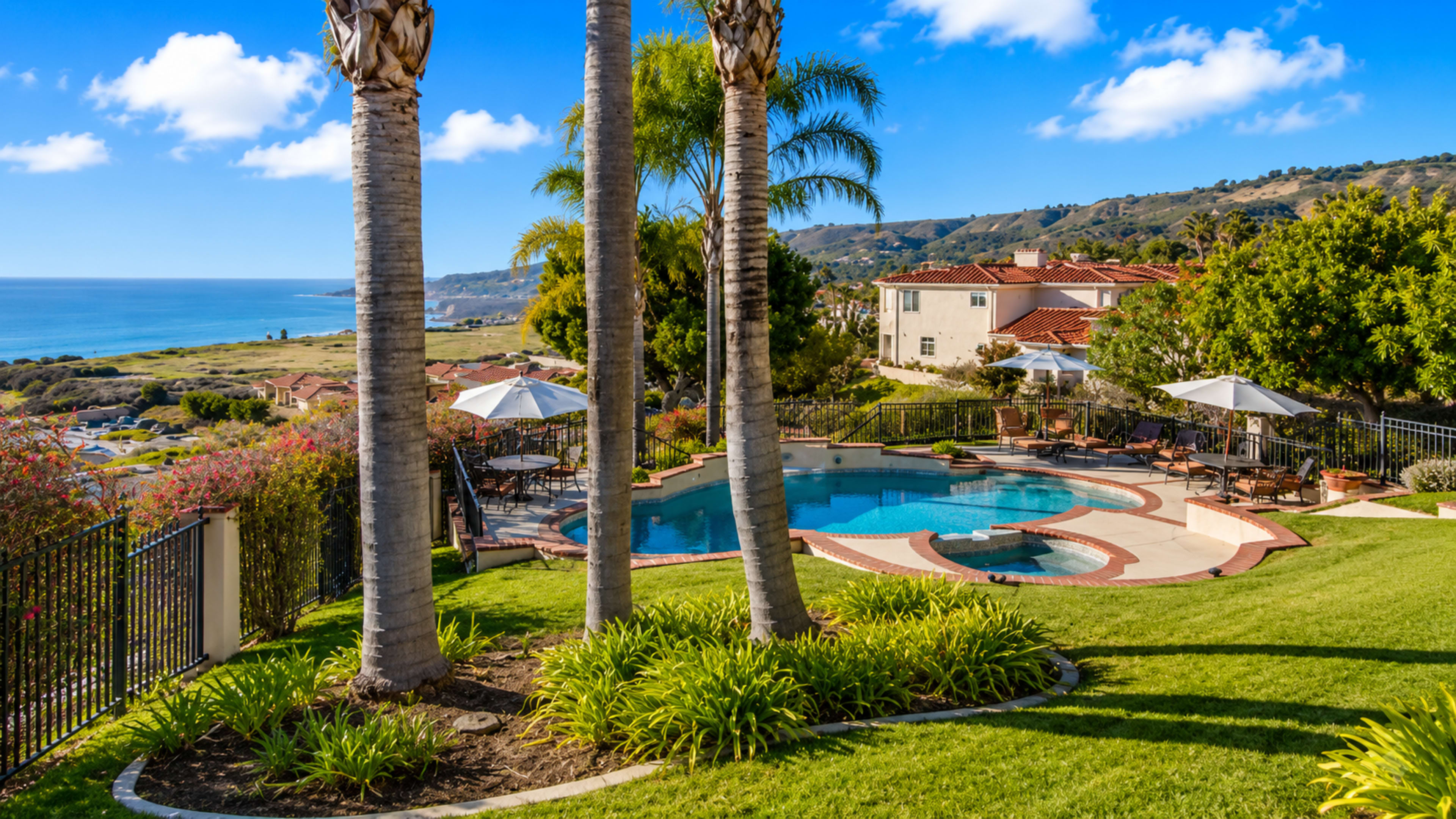 The image depicts a view of a hillside property featuring a swimming pool surrounded by palm trees, with a coastal landscape in the background.