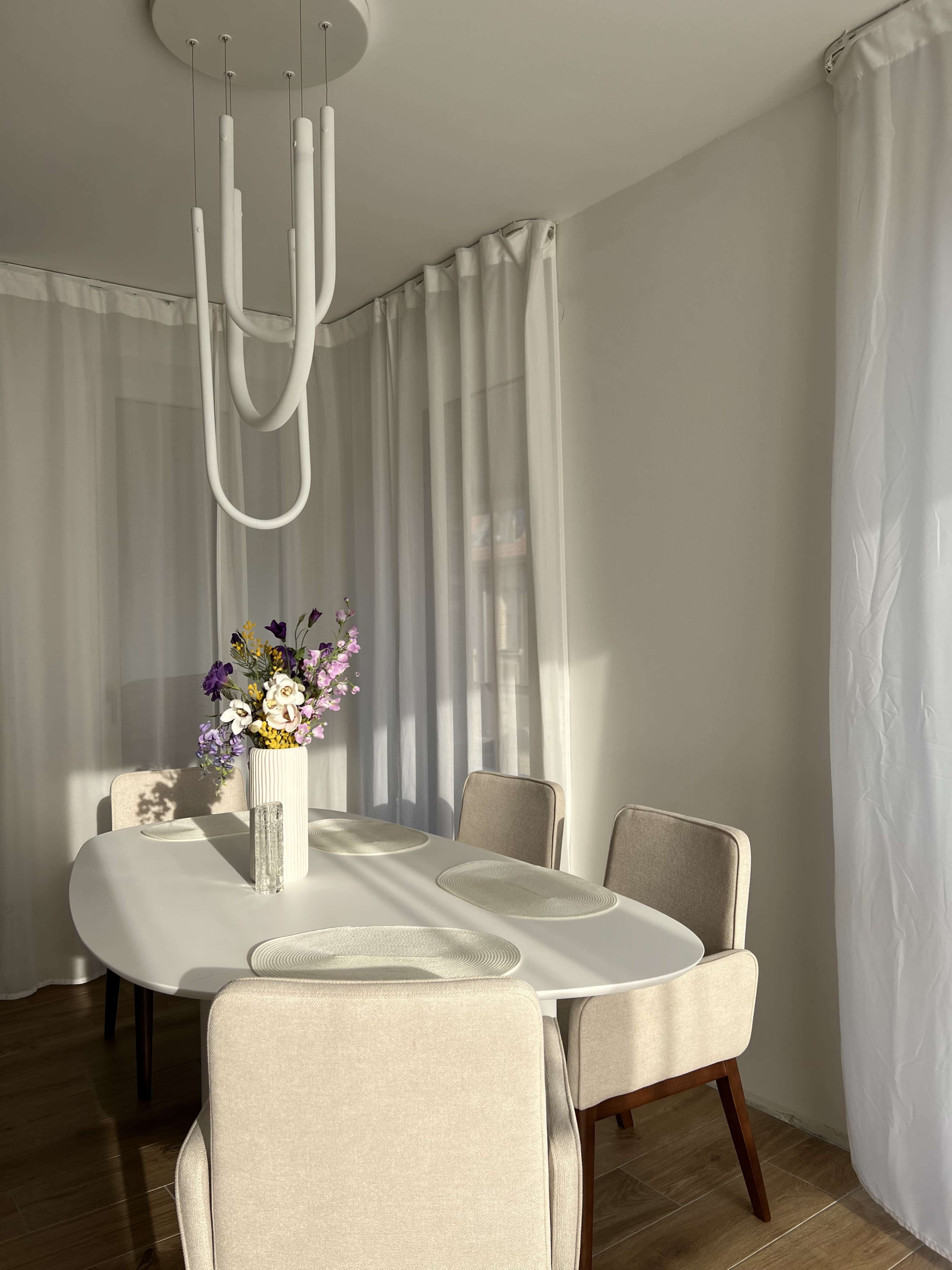A modern dining area features a white oval table with beige chairs, a floral centerpiece, and large sheer curtains.