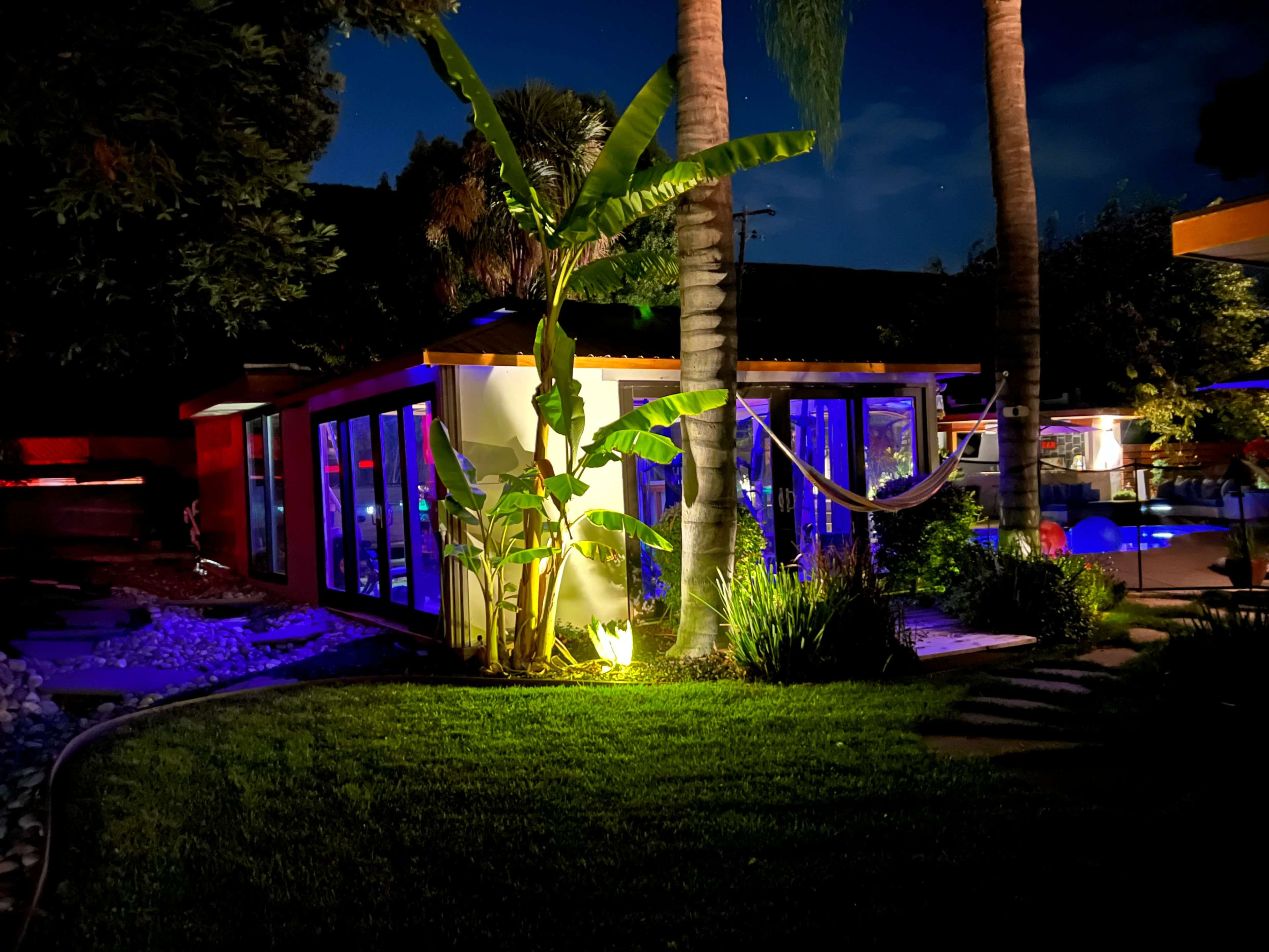 A modern house with large glass windows is illuminated by colorful lights at night, surrounded by palm trees and a well-kept lawn.
