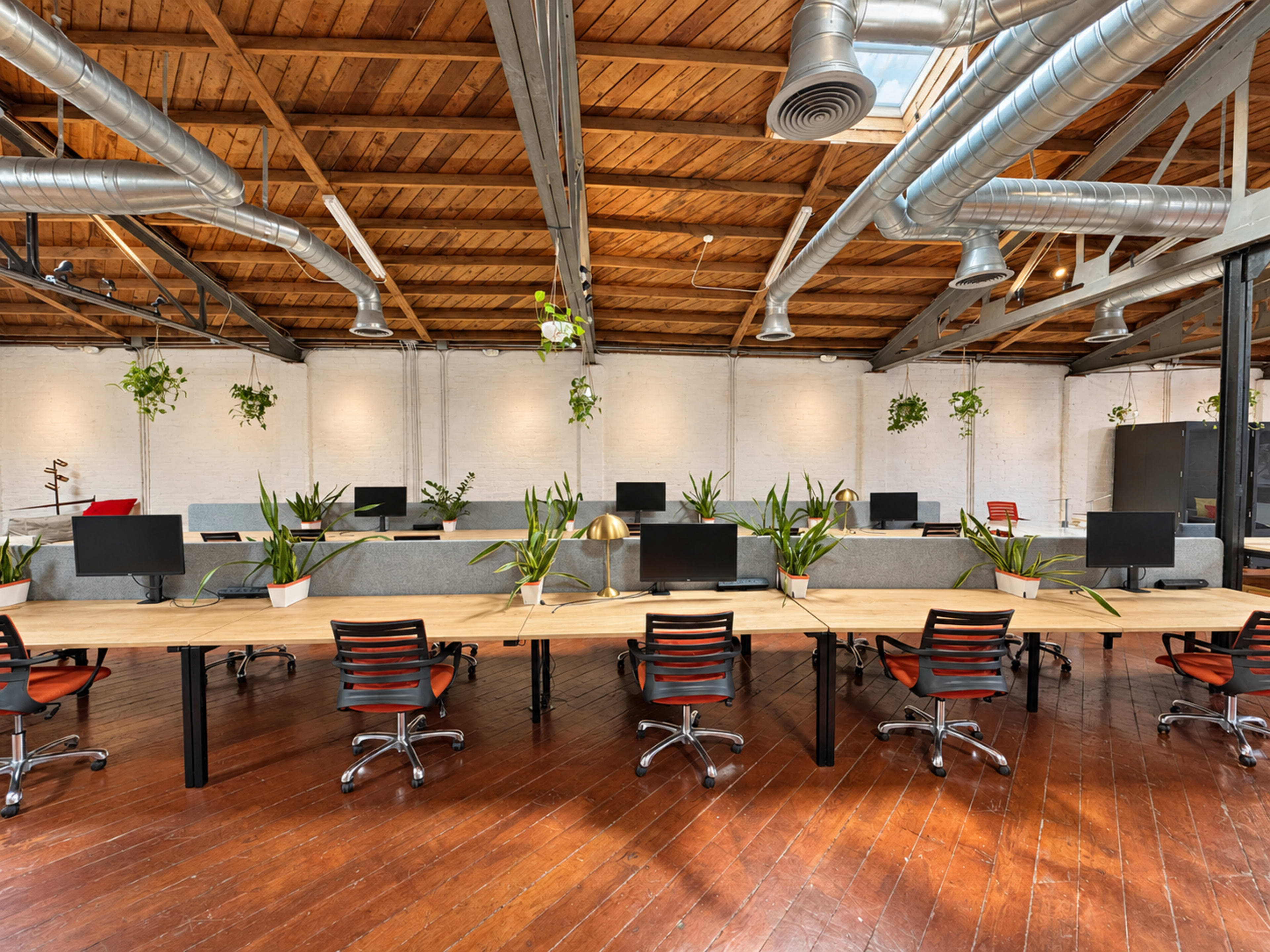 The image shows a spacious, modern office environment with wooden floors, long work tables equipped with computers, and numerous potted plants arranged throughout the space.