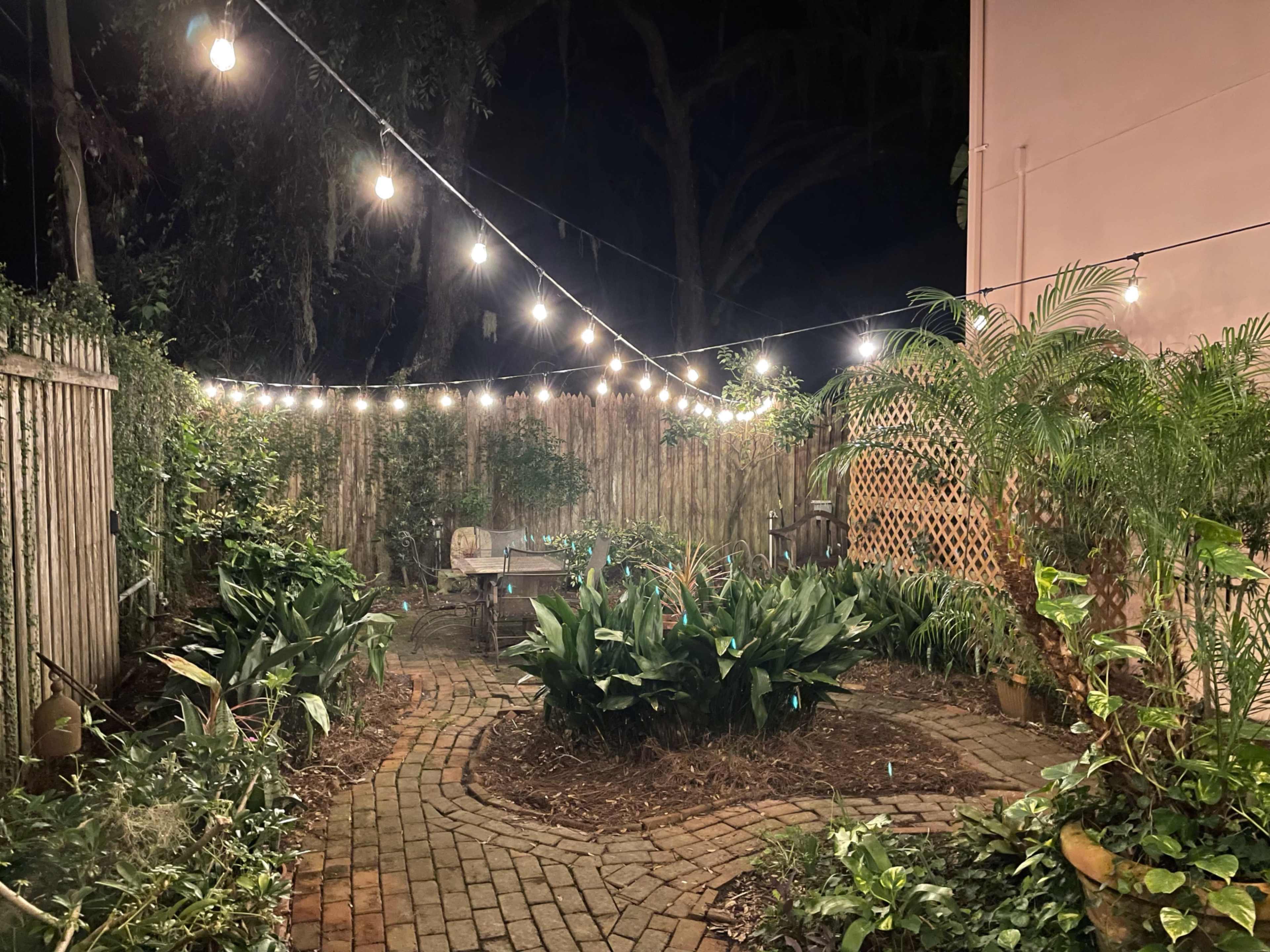 The scene shows a brick path winding through a garden illuminated by string lights at night.