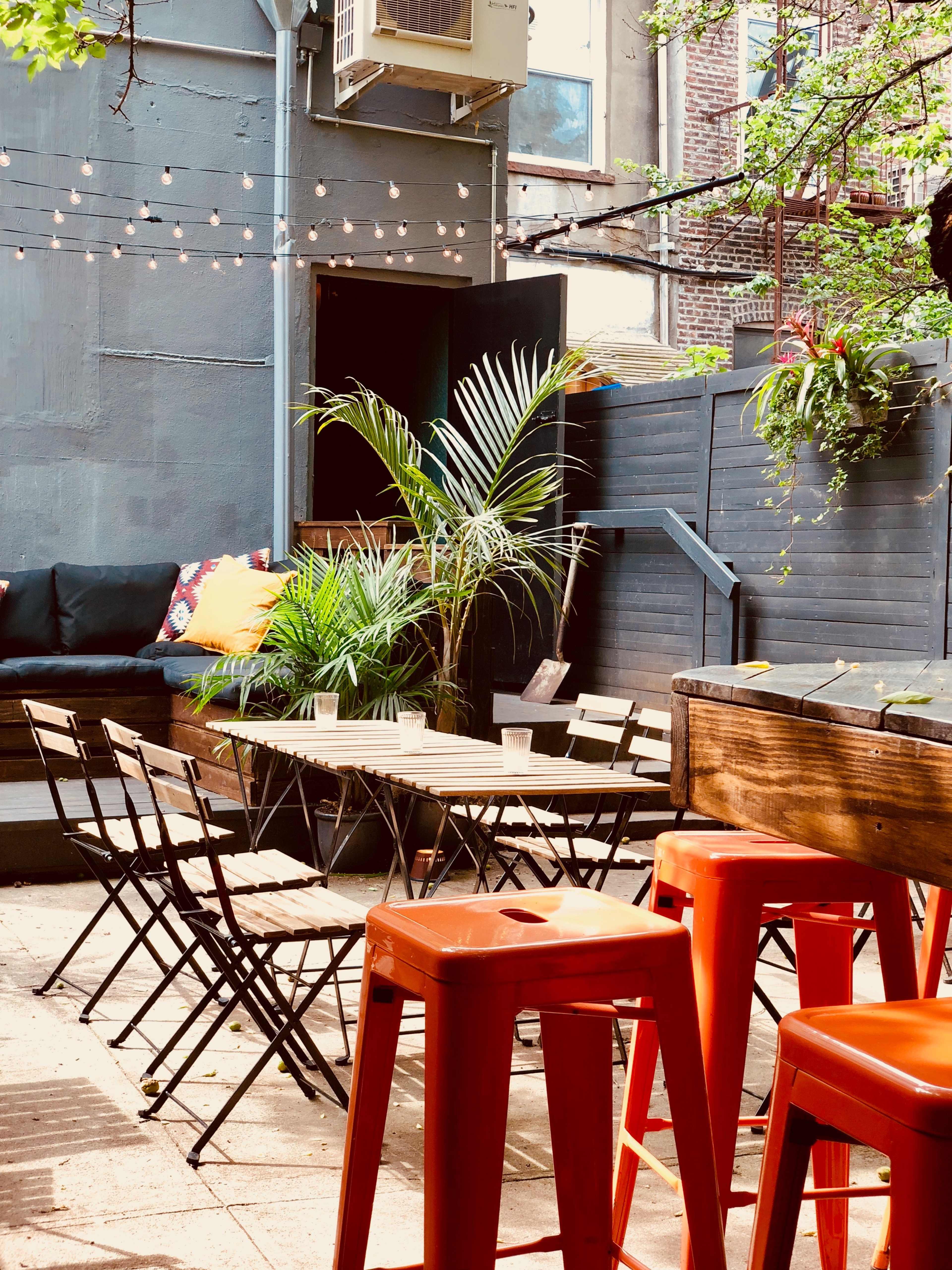 The image shows a small outdoor seating area with metal chairs and tables surrounded by greenery and string lights overhead.