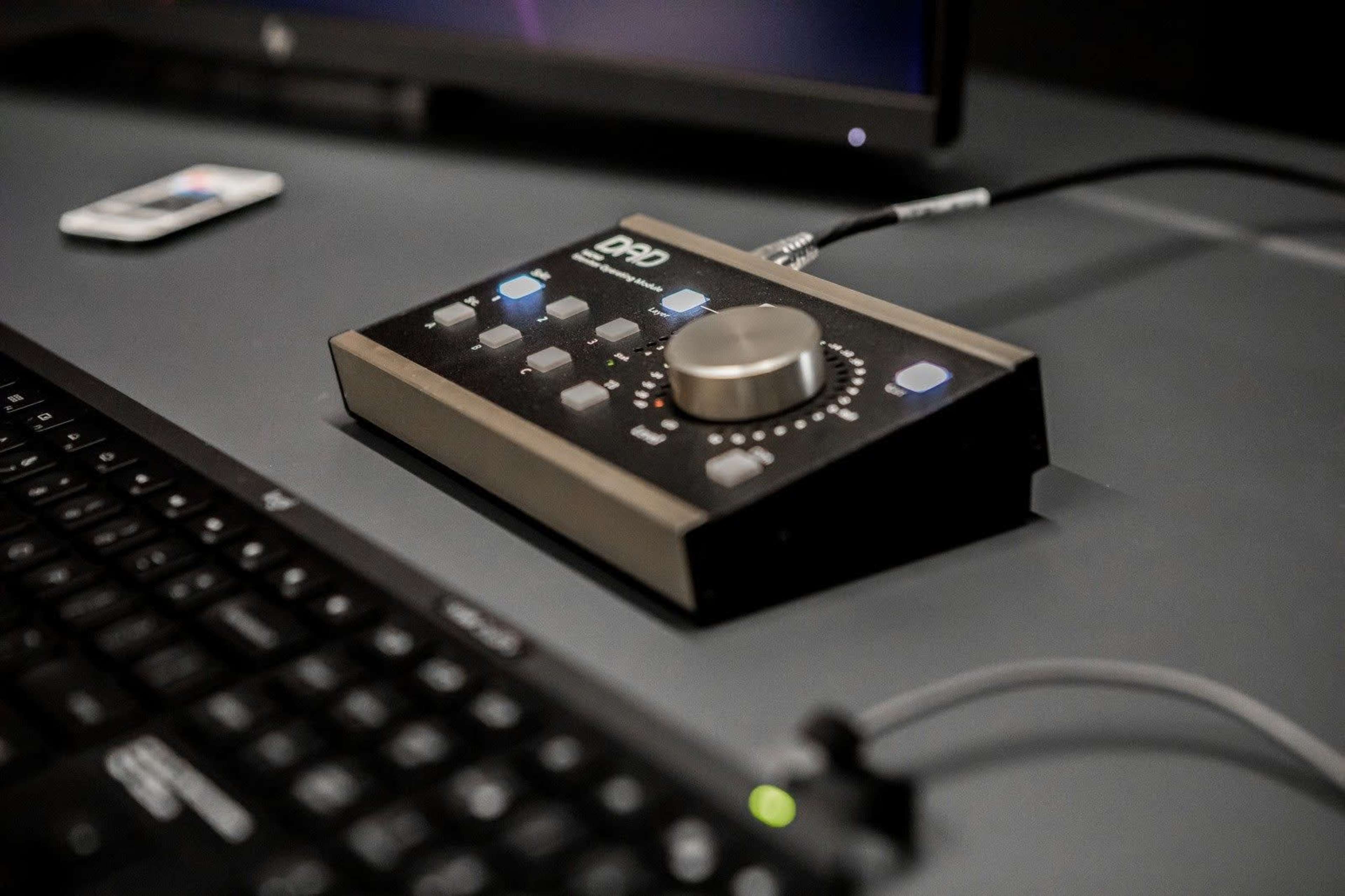 A compact audio interface with knobs and buttons is positioned next to a keyboard on a desk.