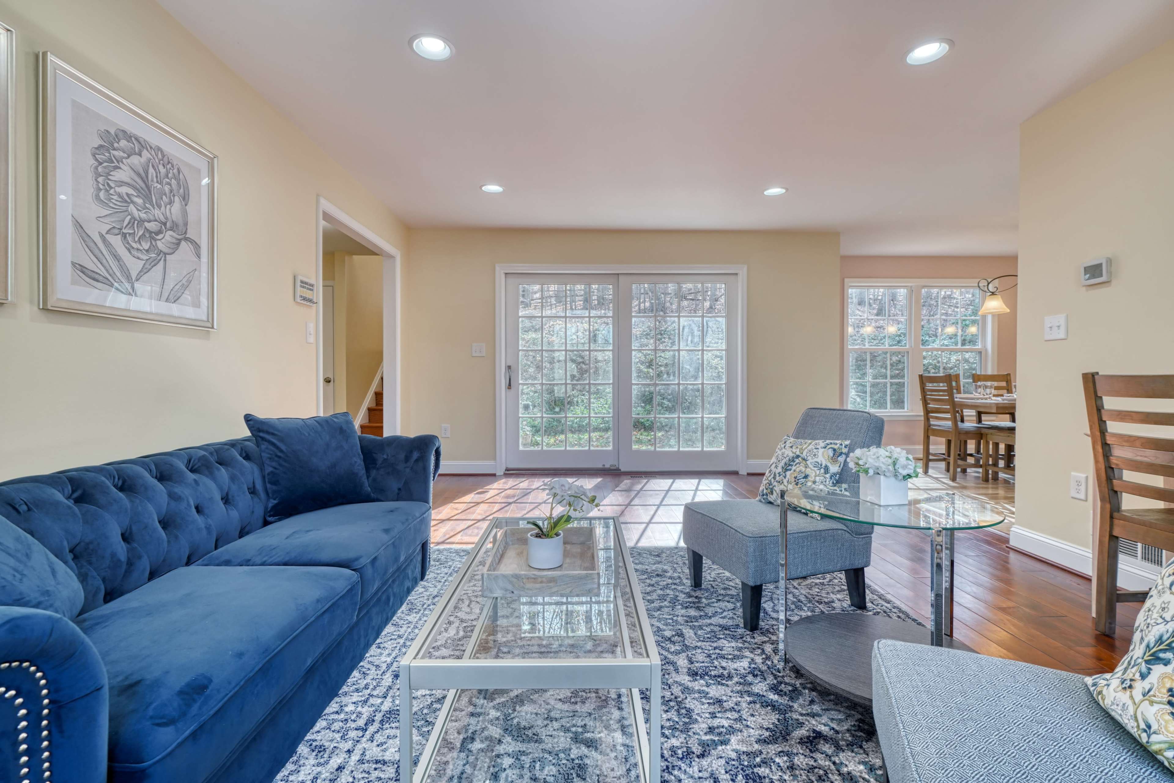 A living room features a blue velvet sofa and two gray chairs, with a glass coffee table on a patterned rug, and large windows allowing natural light to fill the space.