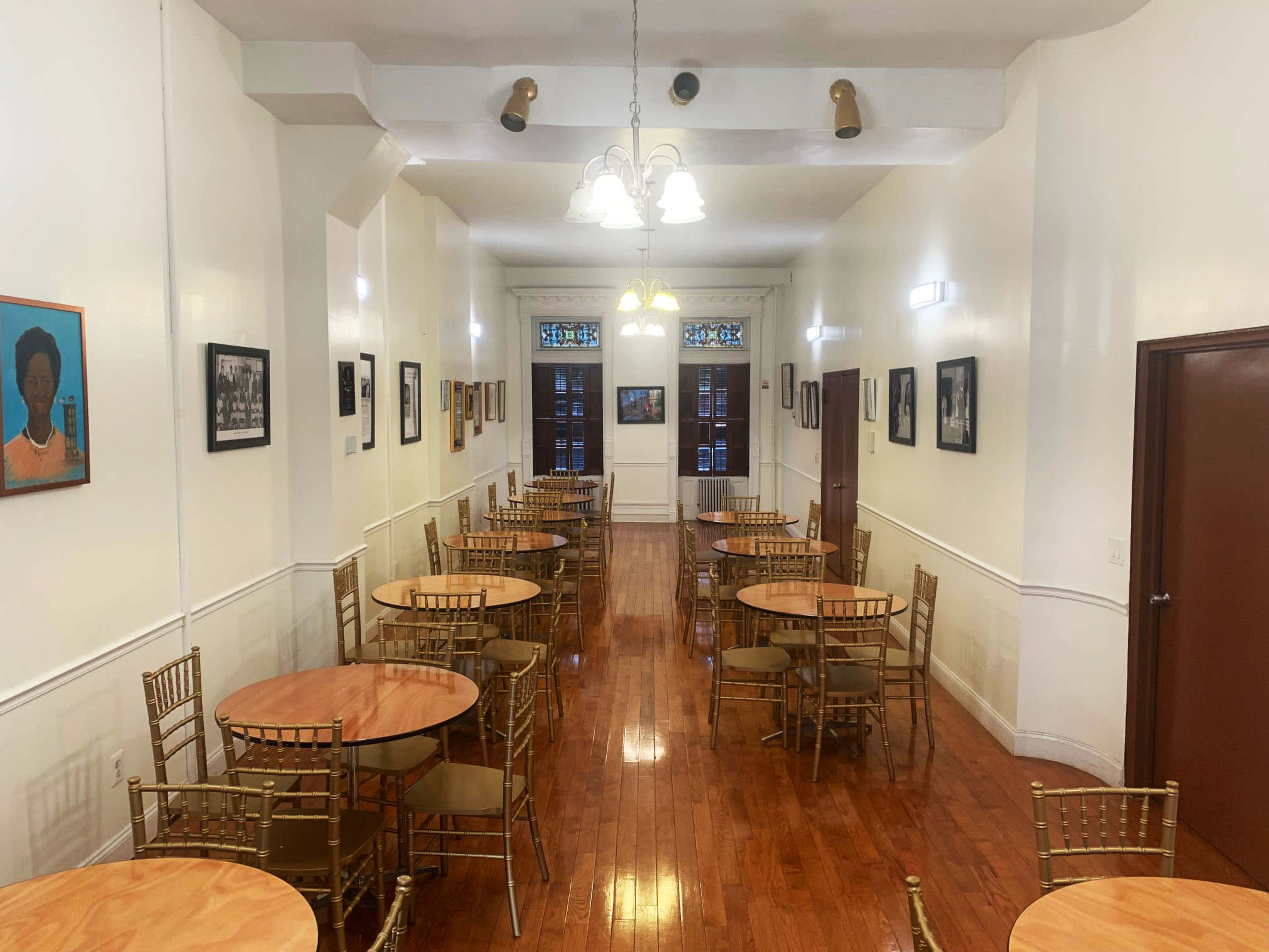 The image shows a long, narrow dining hall with several round wooden tables surrounded by gold-colored chairs, and walls adorned with framed photographs and artwork.