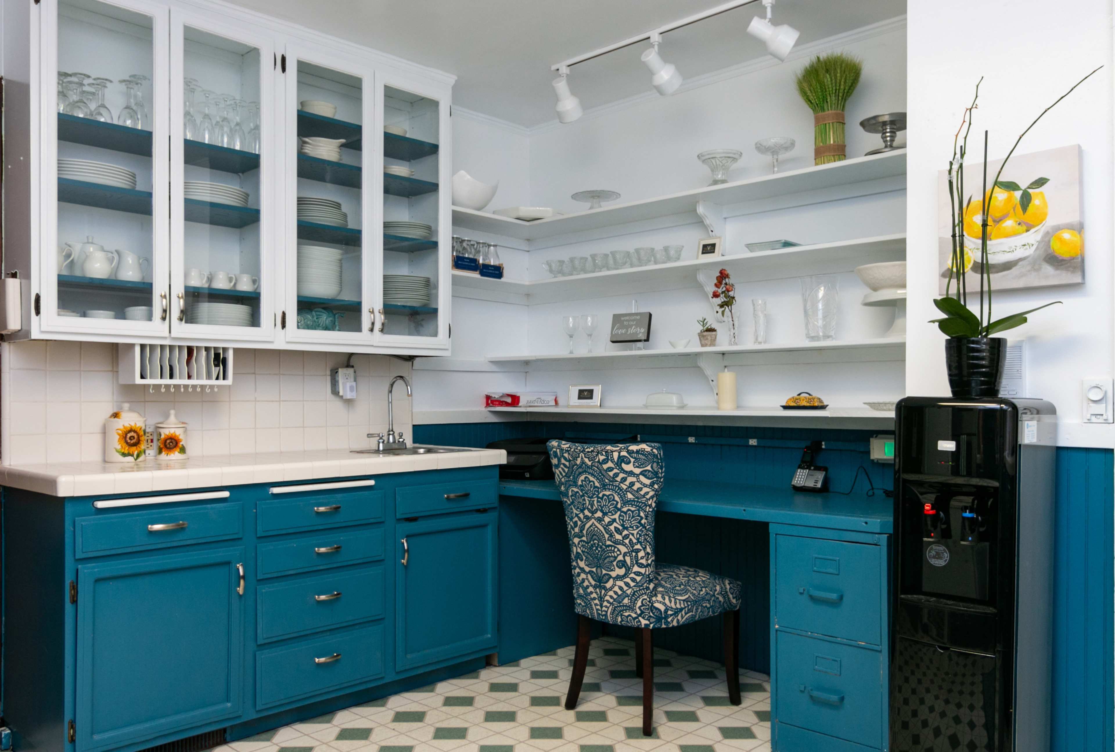 A modern kitchen with blue cabinetry, a desk area, and glass-front upper cabinets displaying dishware.