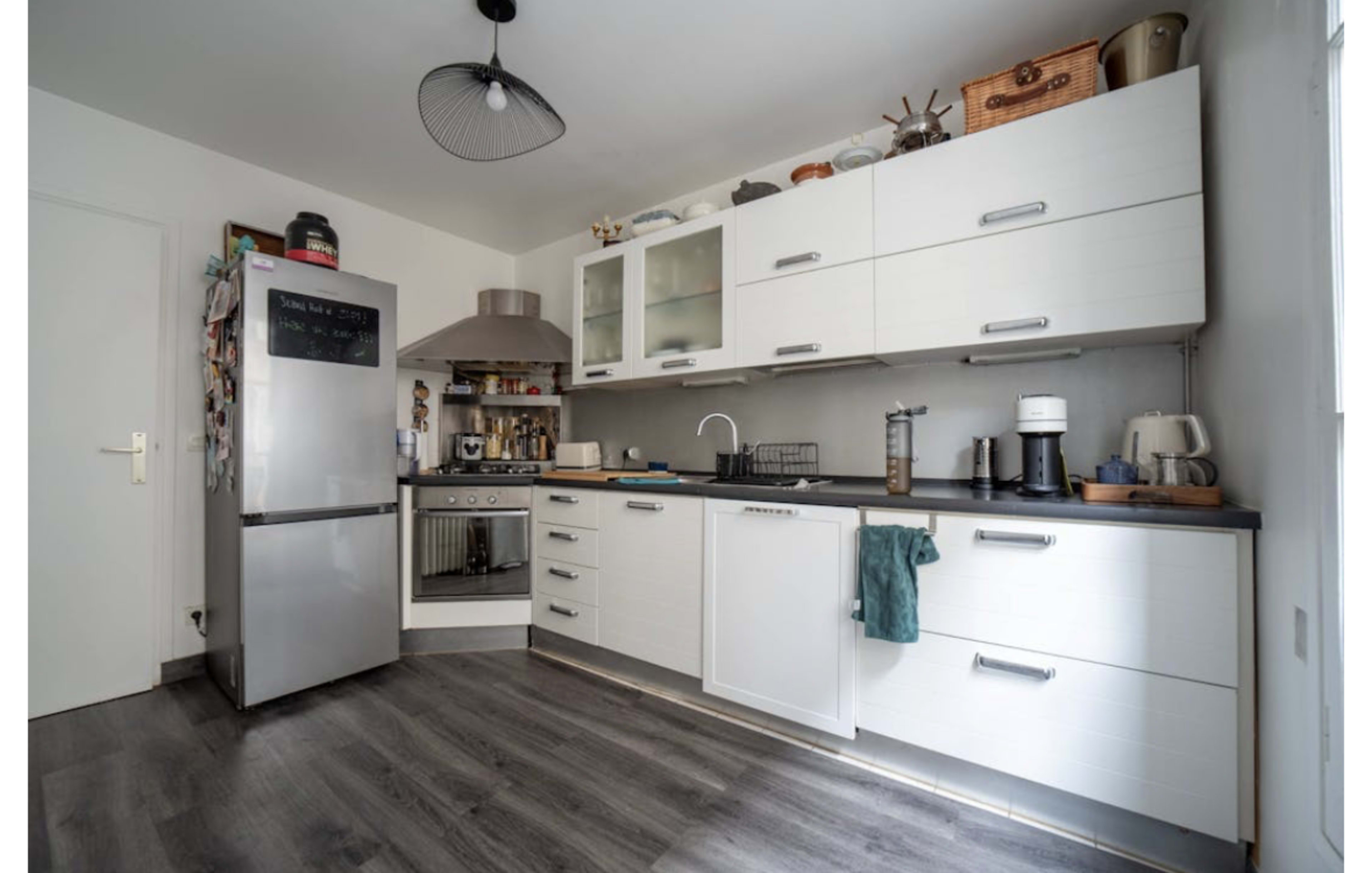 The image shows a modern kitchen with white cabinets, a stainless steel refrigerator, and a dark countertop, featuring an oven and various kitchen appliances.