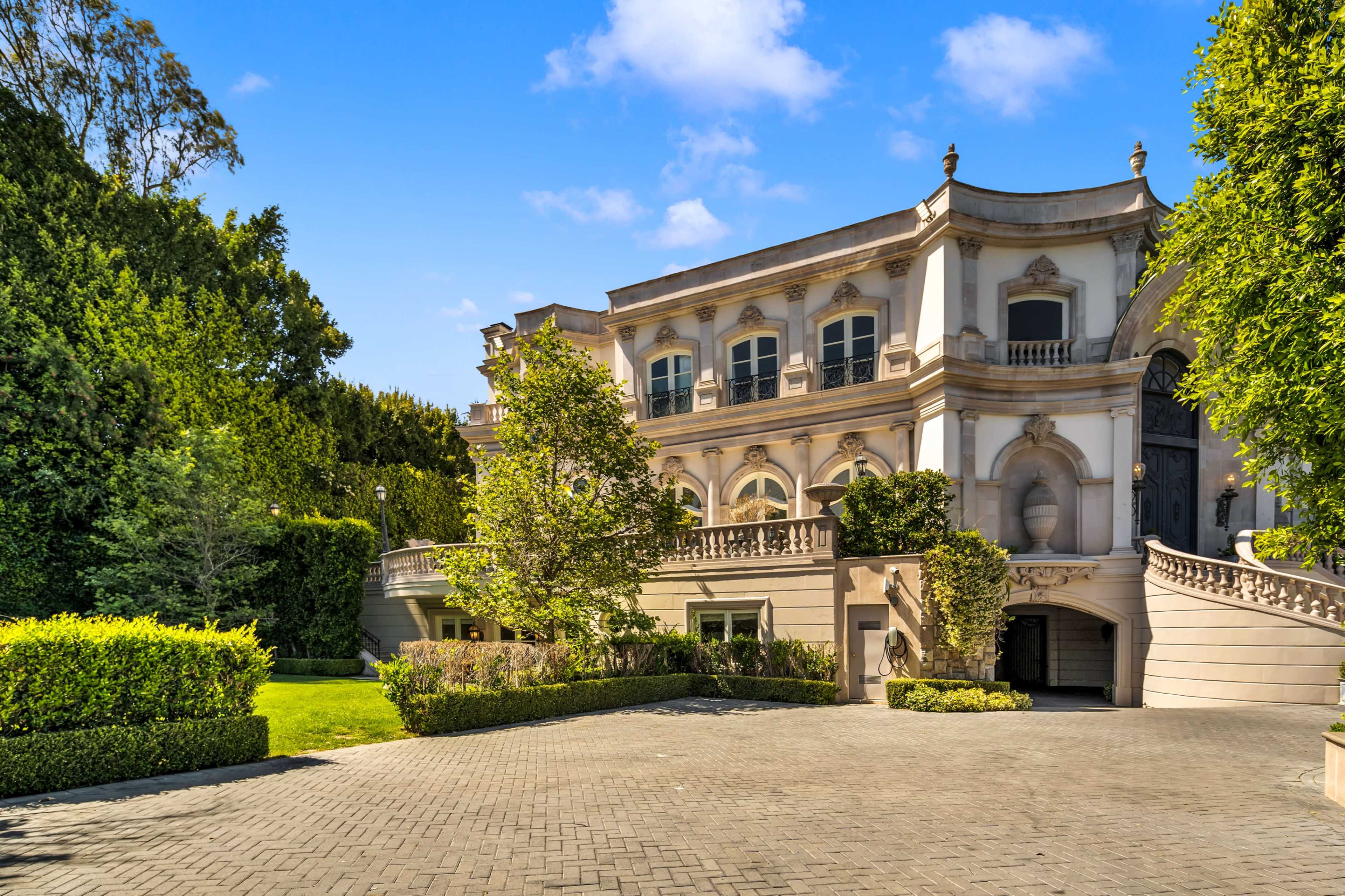 A large, ornate mansion with a well-maintained garden and a paved driveway is set against a clear blue sky.