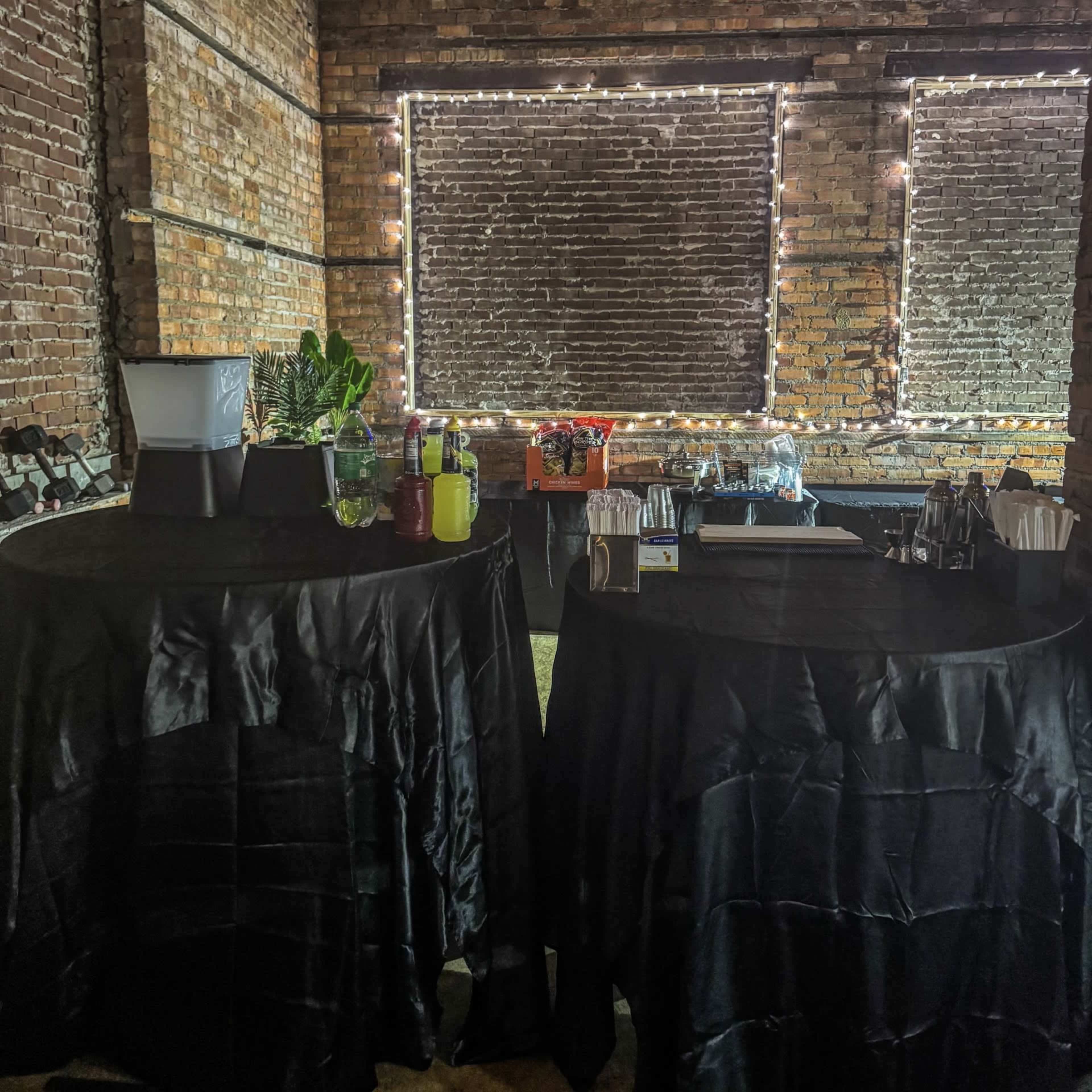 Two black-cloth-covered tables are set up in a brick-walled room, displaying various drinks and supplies under string lights.