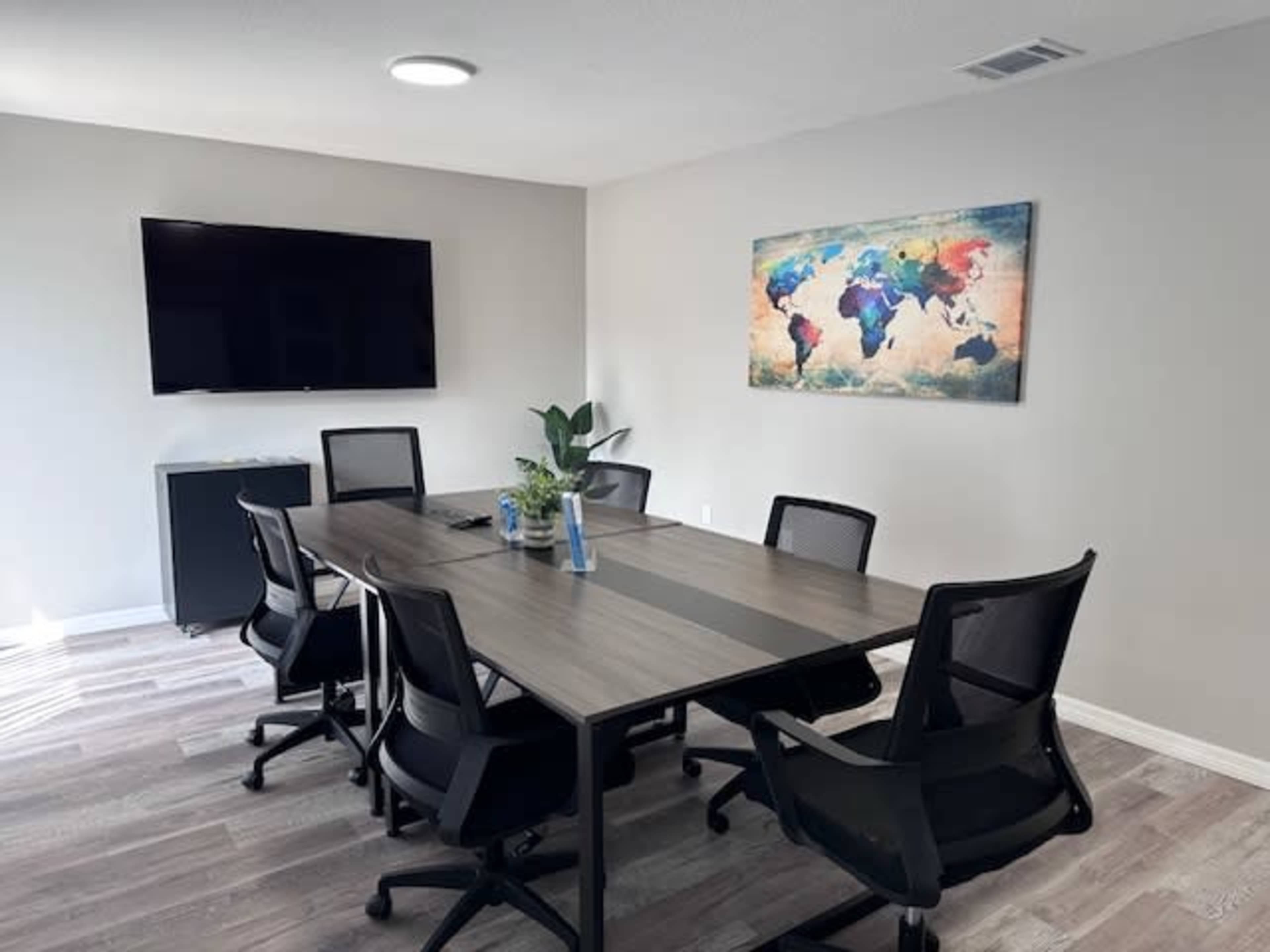 A modern conference room features a large table surrounded by six black office chairs, a wall-mounted TV, and a colorful world map artwork.