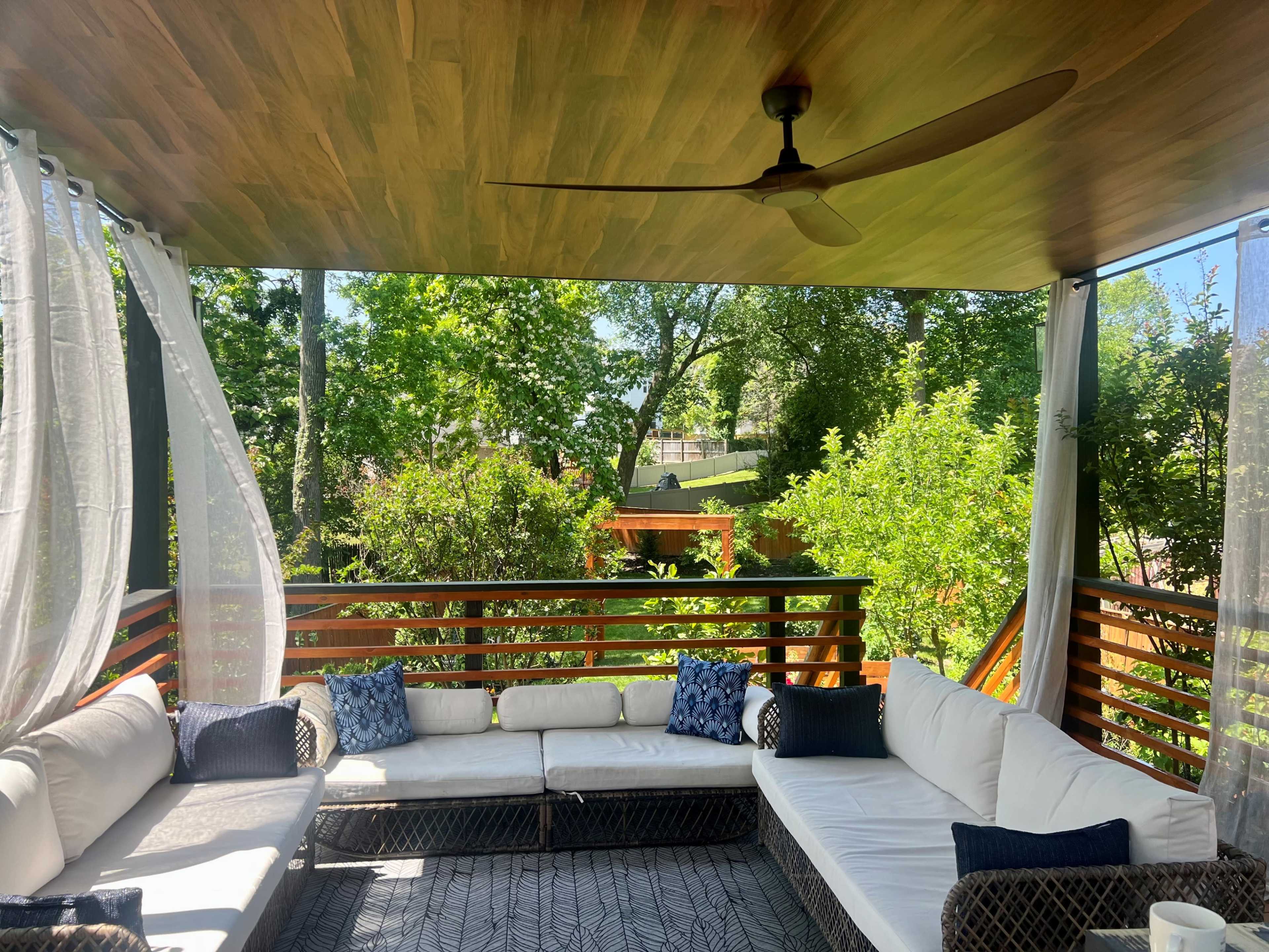 A covered patio with a sectional sofa, blue decorative pillows, and sheer curtains overlooks a garden filled with greenery.