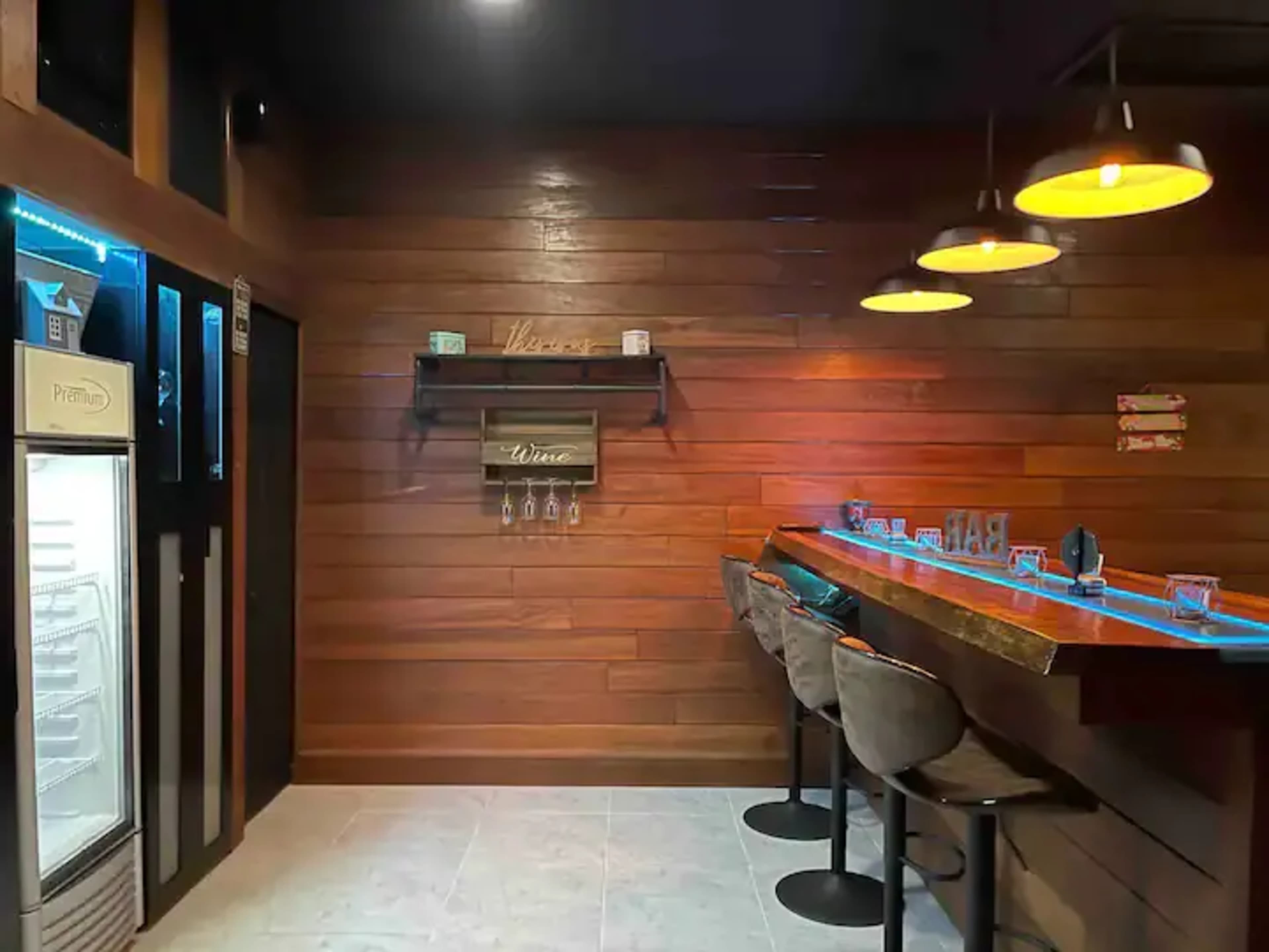 A modern bar area with wooden paneling, four stools arranged at a countertop, and a refrigerator in the corner.