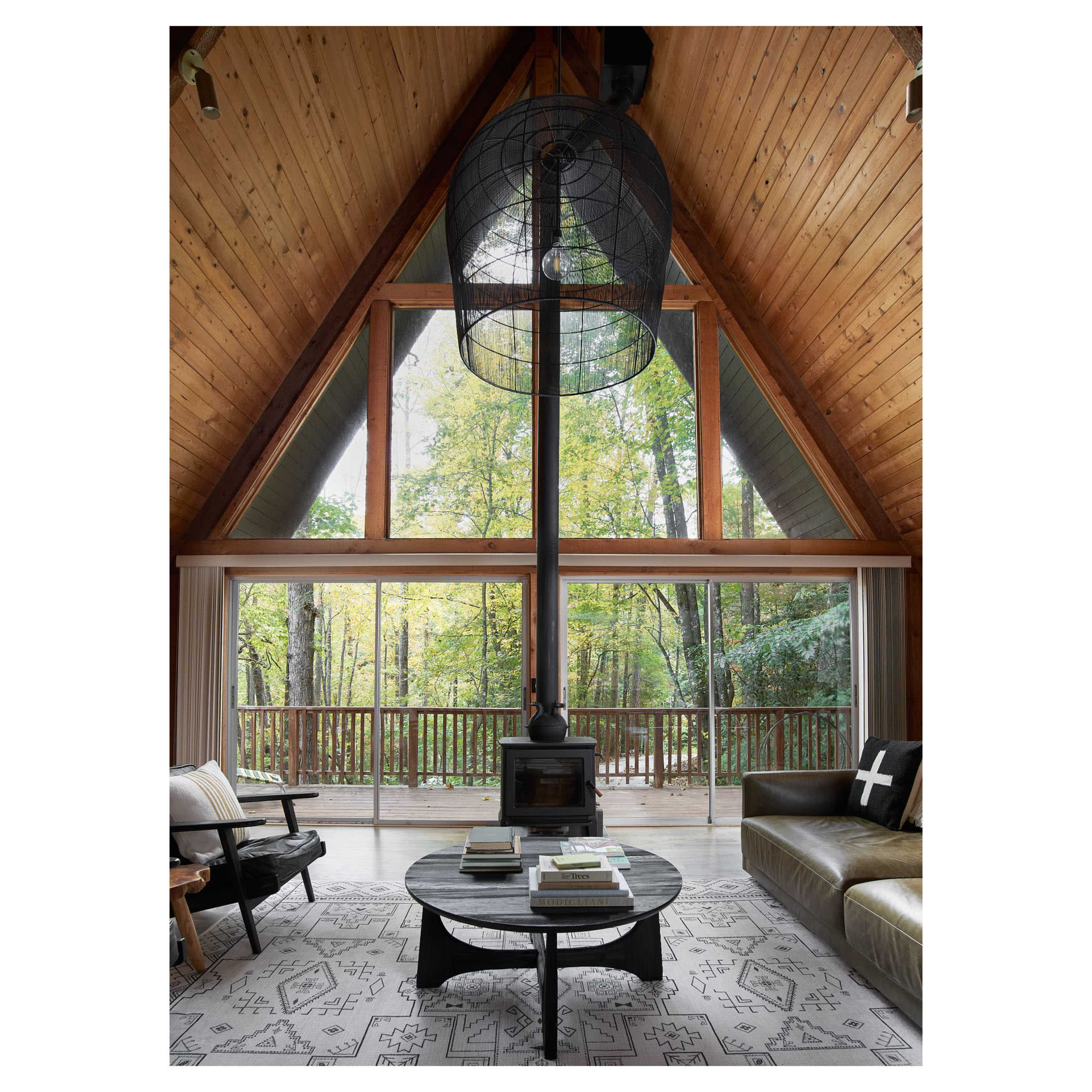 A modern cabin interior with a high wooden ceiling, large windows overlooking a forest, and a circular coffee table arranged with books.