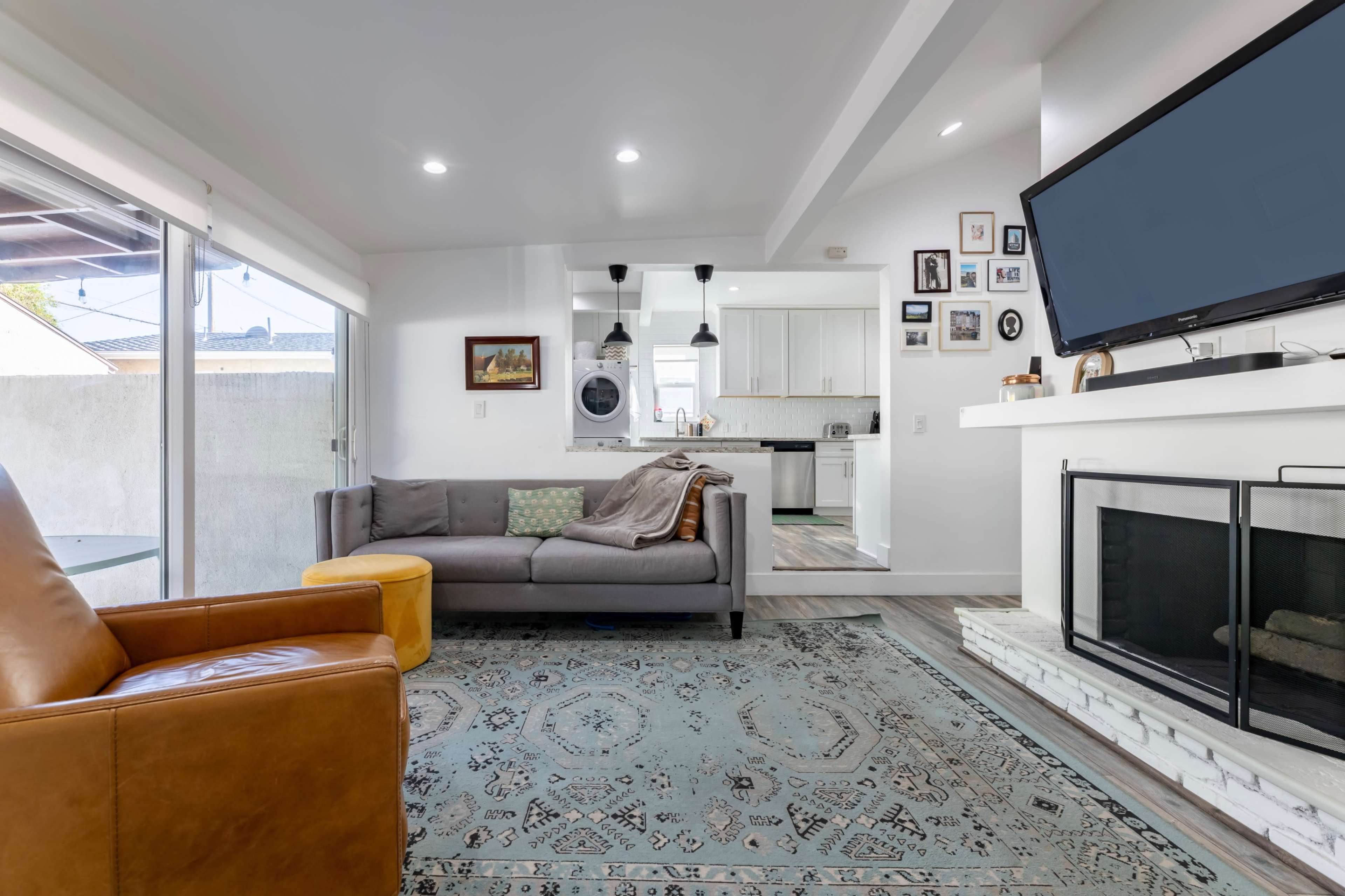 The image shows a modern living room with a gray sofa, a brown leather chair, and a stone fireplace, leading into a kitchen area.