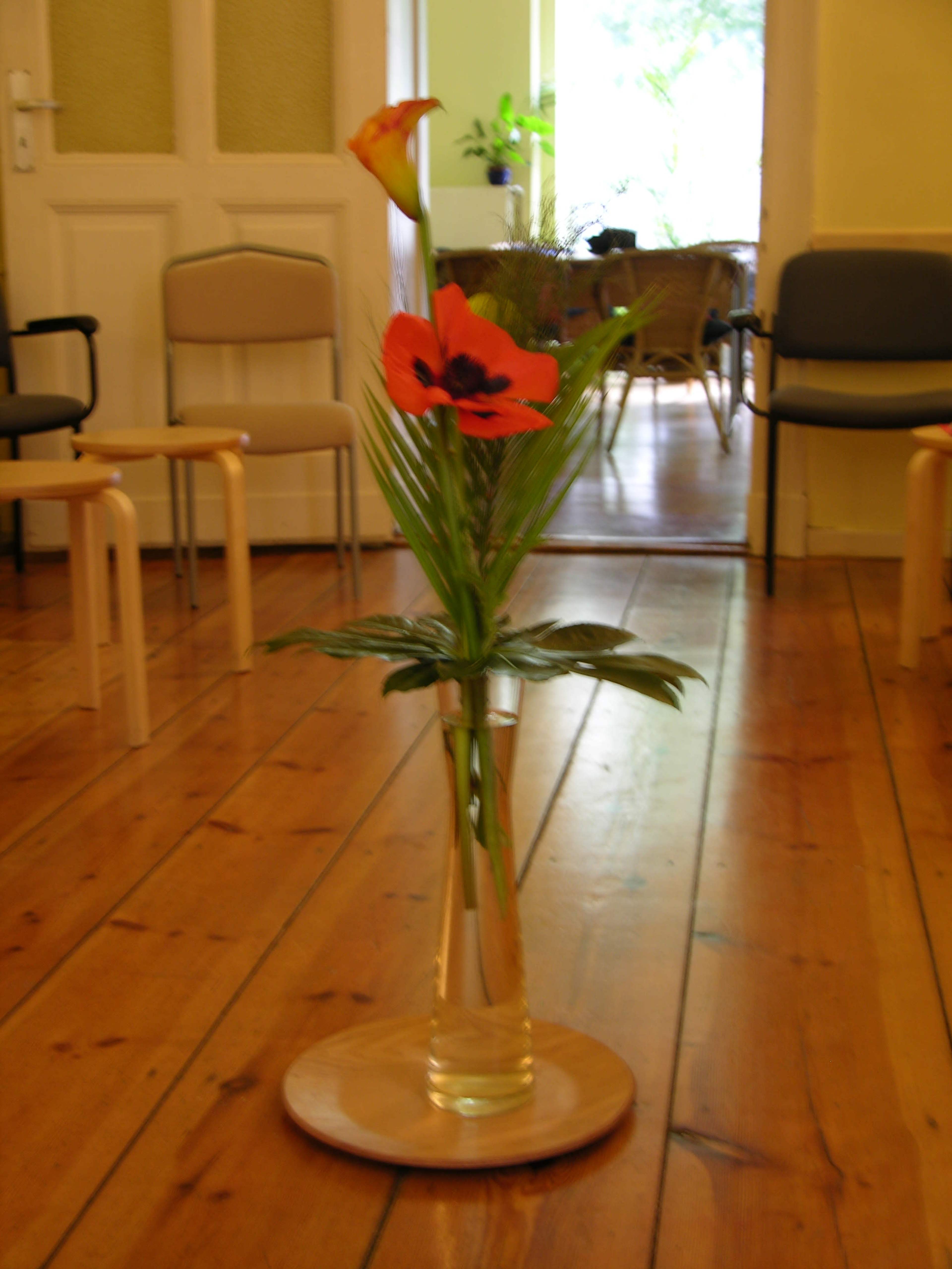 A glass vase with flowers is displayed on a wooden floor in a room with chairs in the background.