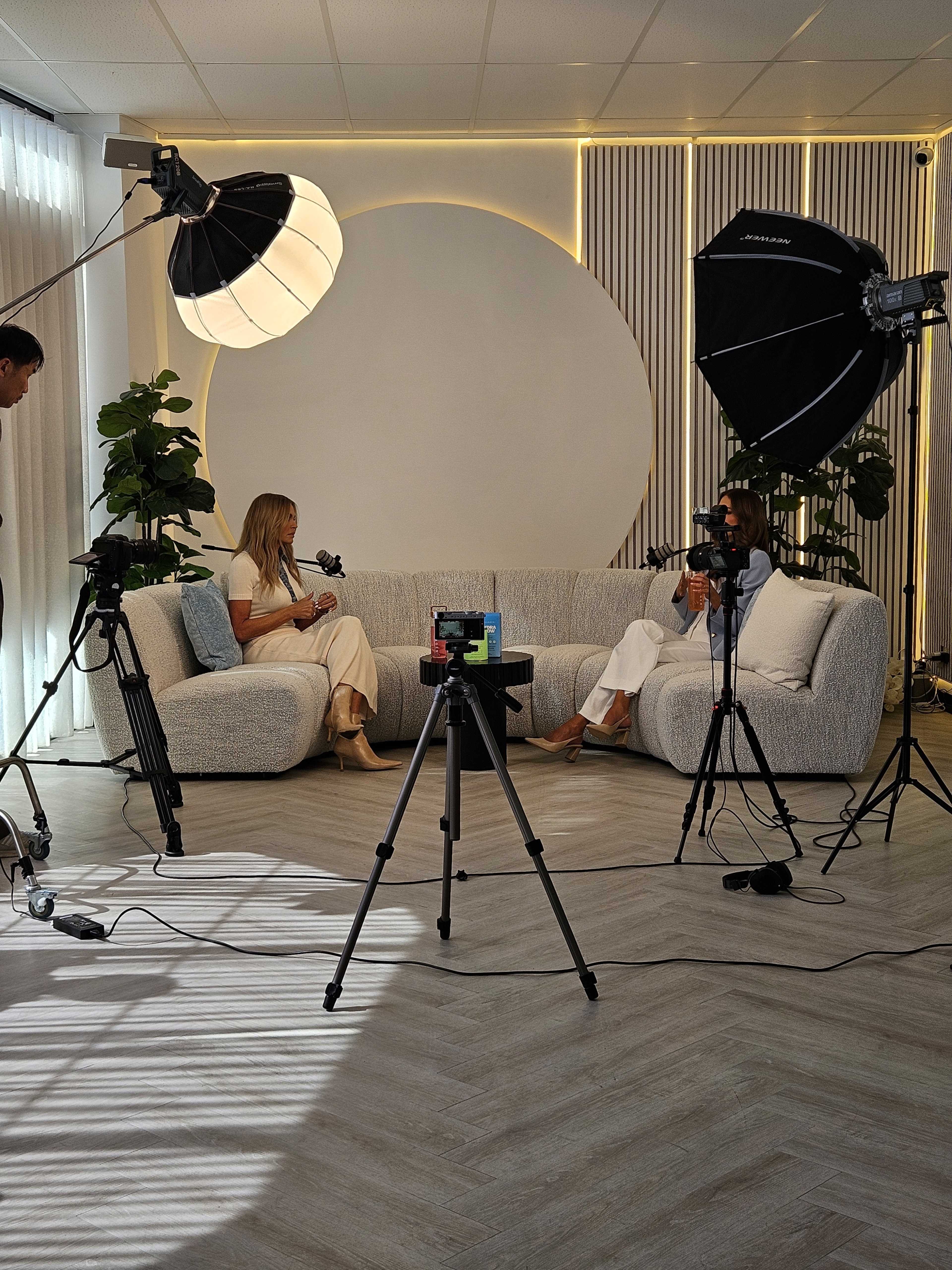 A modern interview setup features two people seated on a light-colored sofa, equipped with professional lighting and cameras in a brightly lit room.