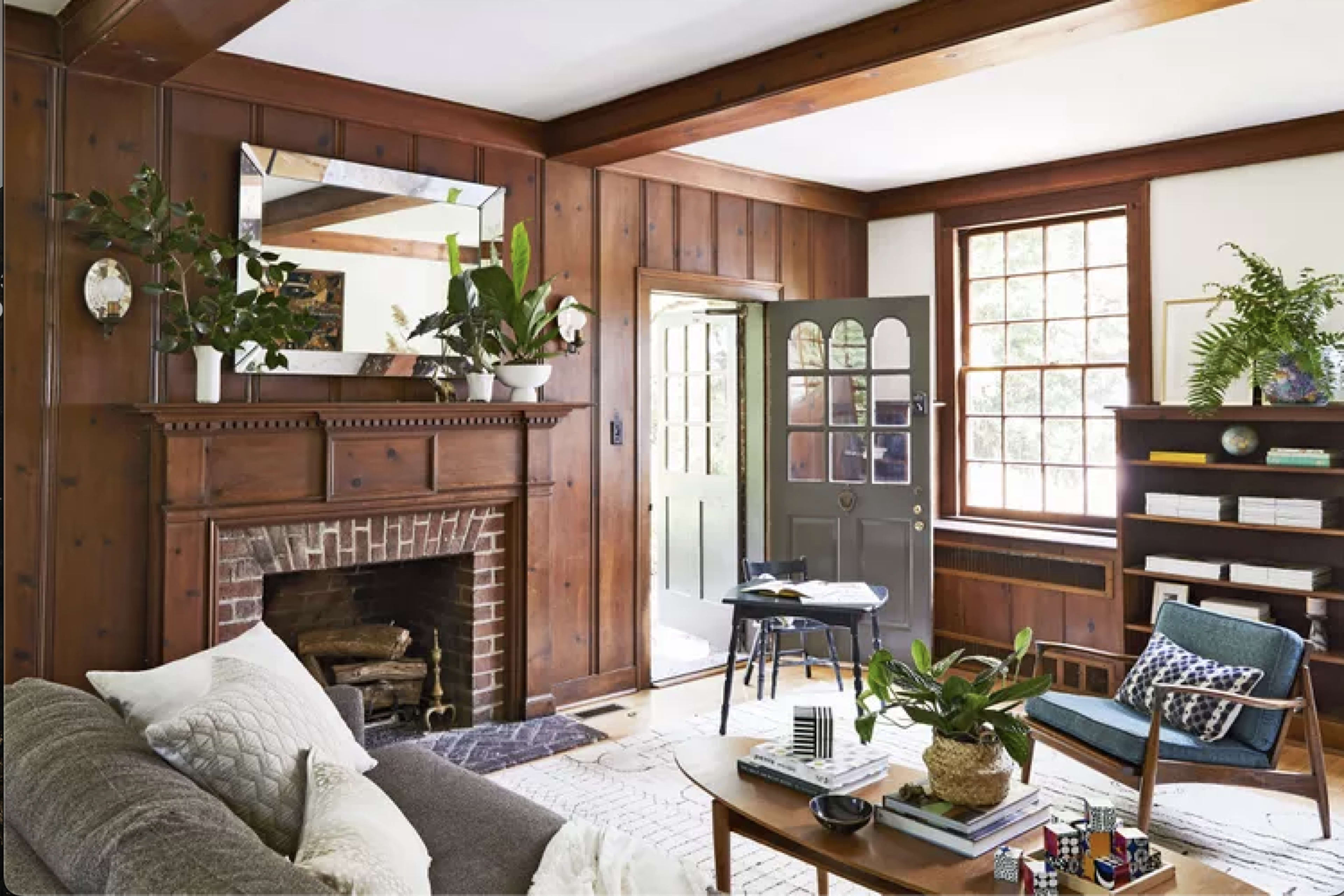 The image shows a cozy living room with wooden walls, a brick fireplace, plants, and a dark door leading outside.