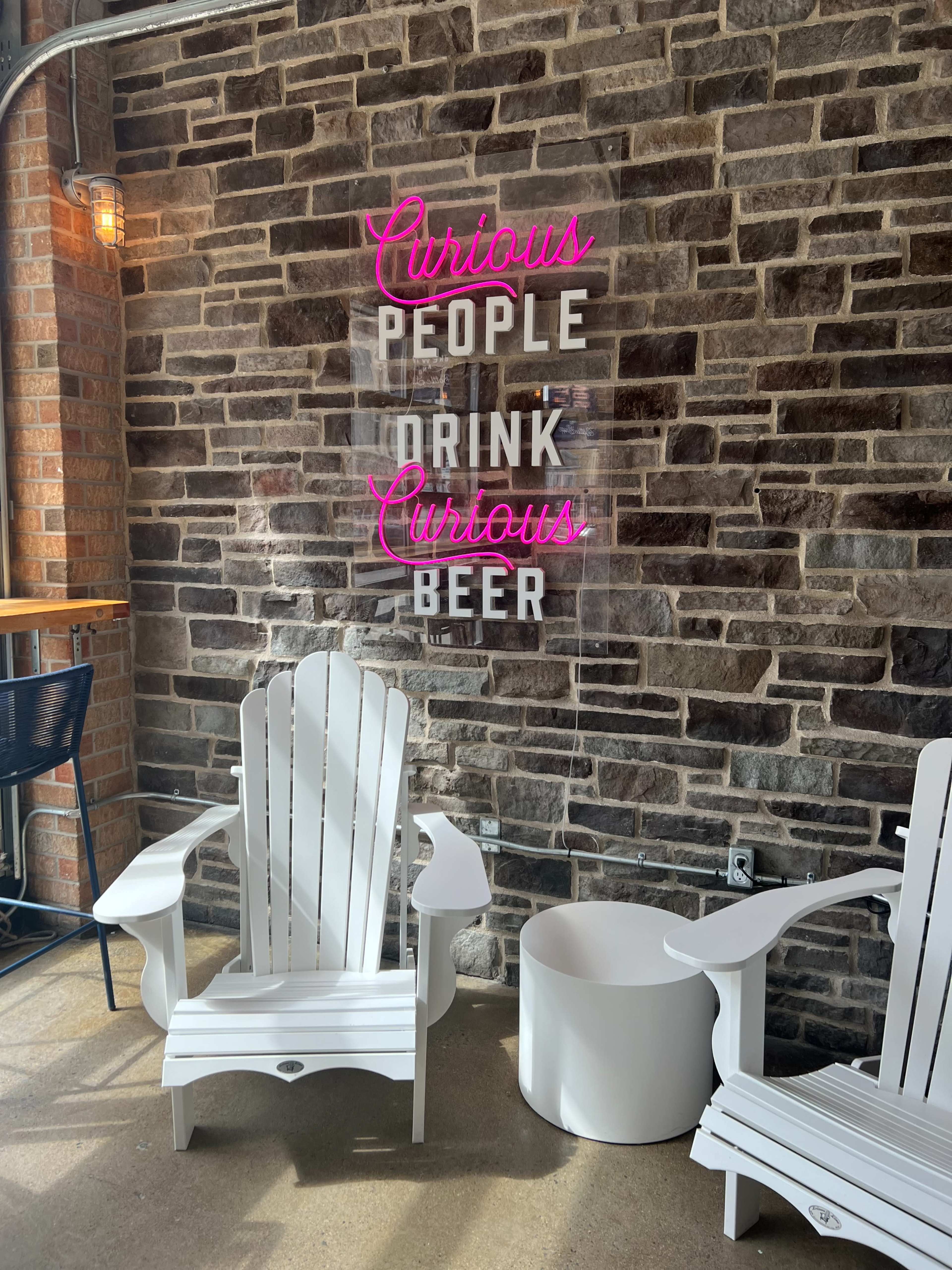 Two white Adirondack chairs in front of a stone wall with a neon sign that reads "Curious People Drink Curious Beer."