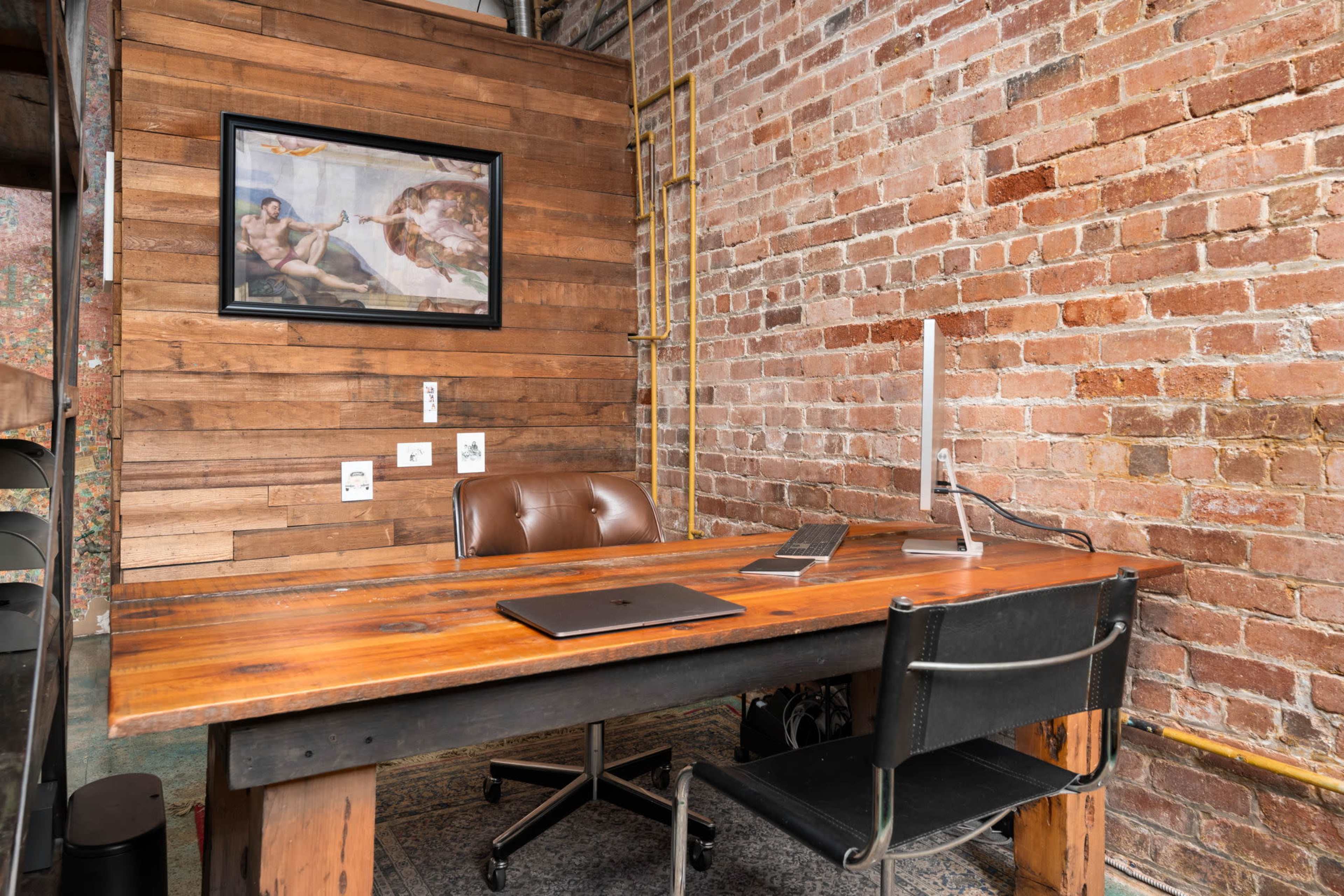 The image shows a rustic office space with a wooden desk, a computer monitor, and a framed artwork on a brick wall.