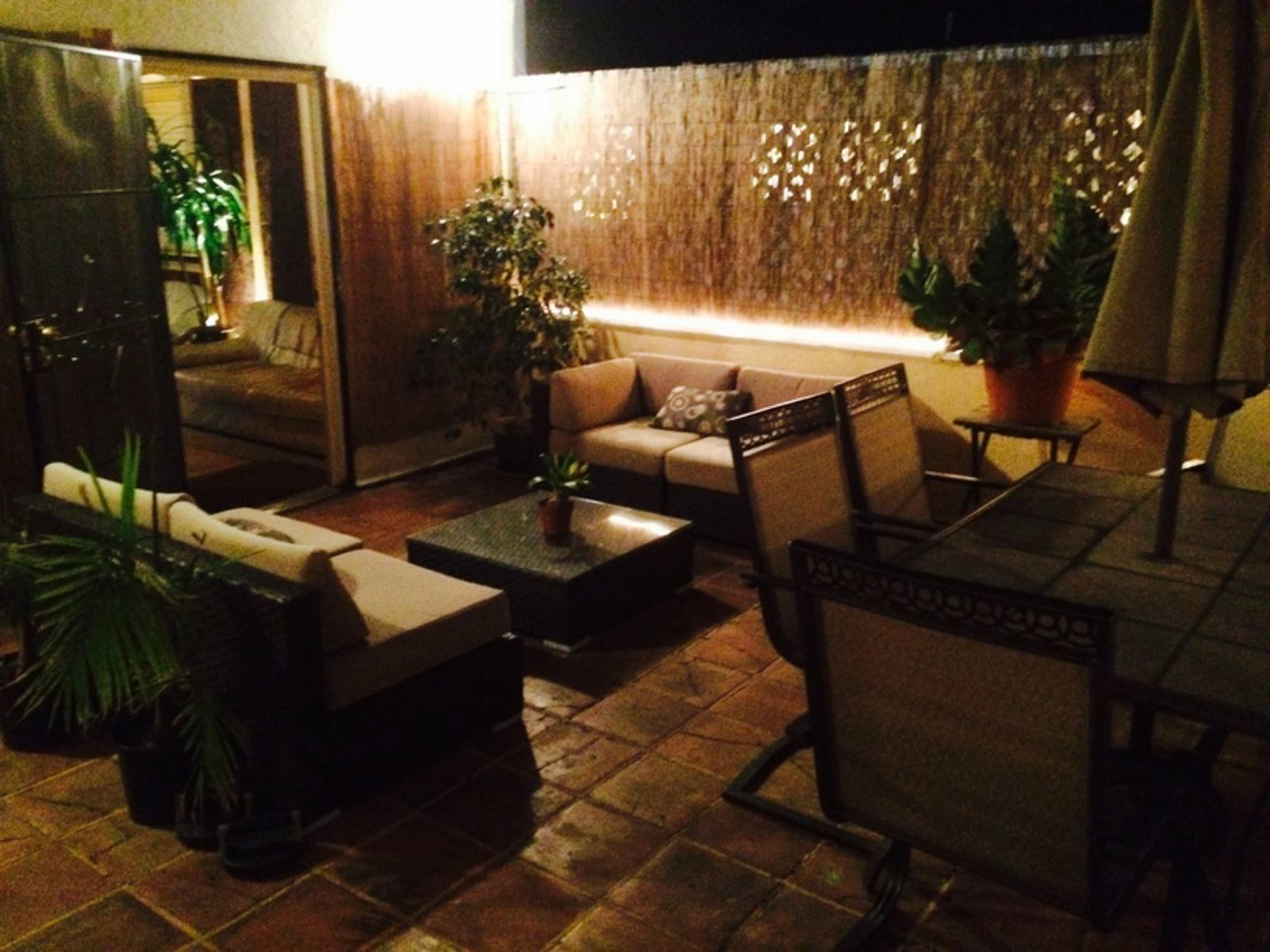 A patio area features a seating arrangement with a coffee table and dining set, surrounded by plants and illuminated by soft lighting.