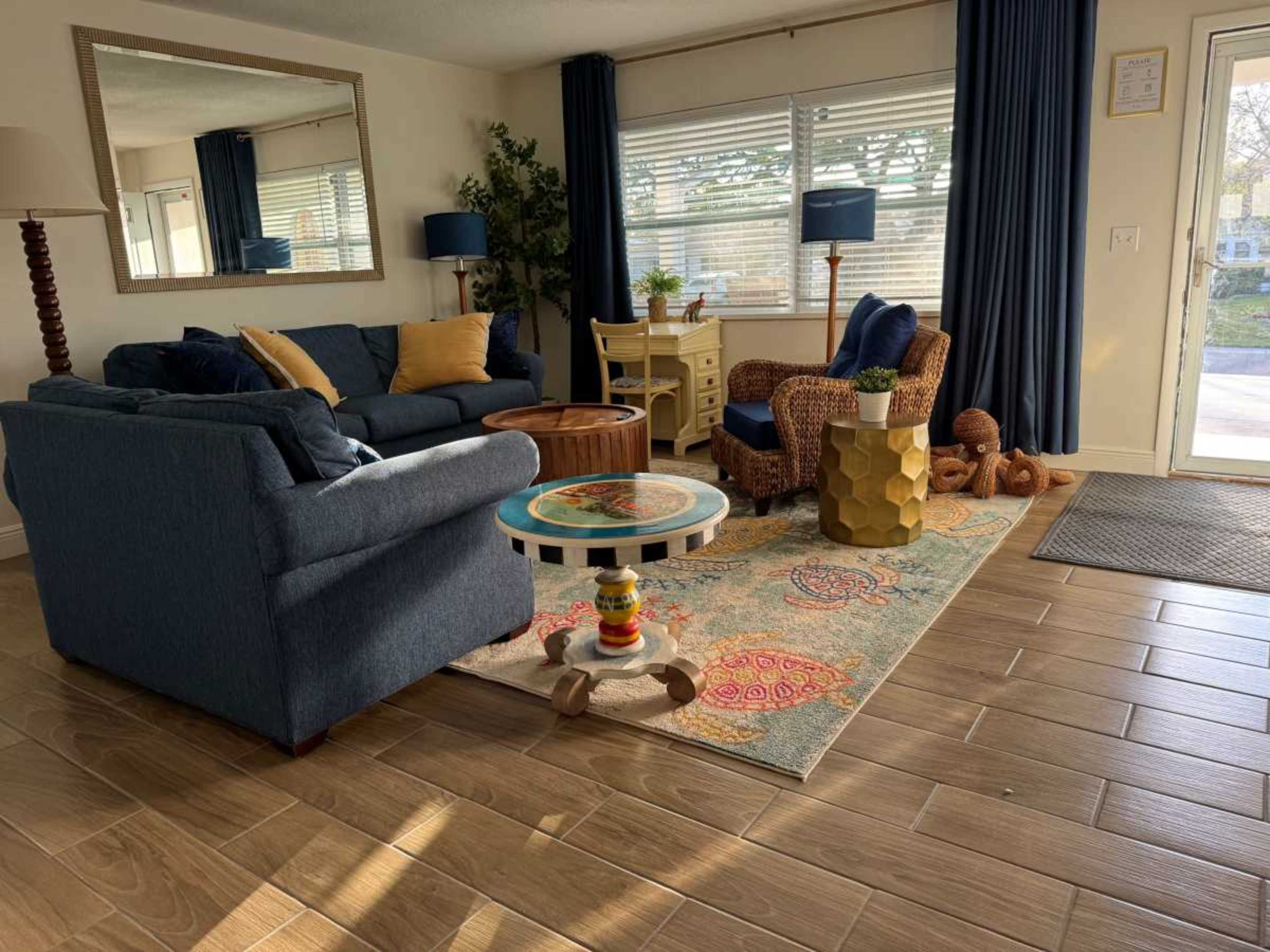 The image shows a bright, inviting living room with a blue sofa, a round coffee table, a patterned rug, and various chairs near a window.