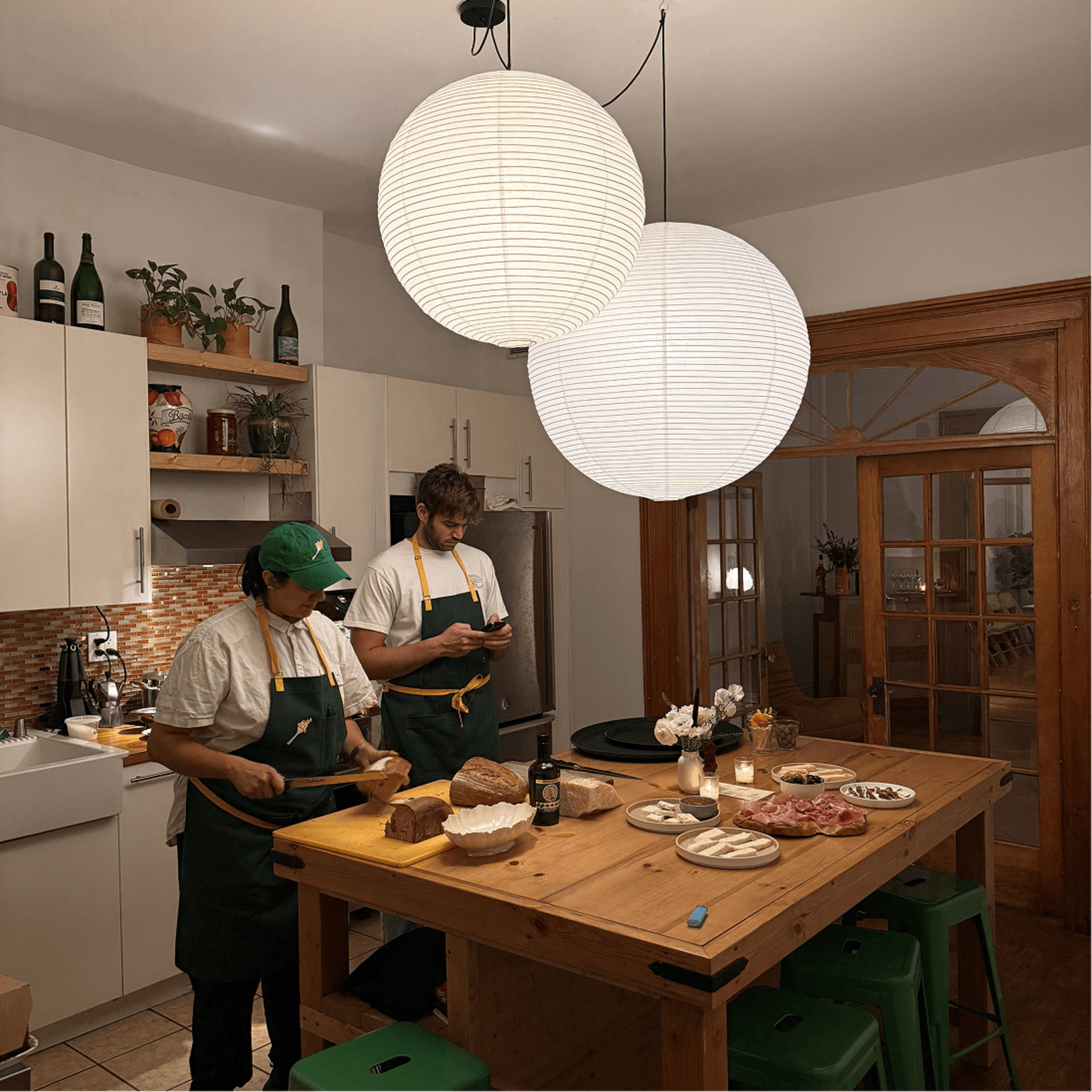 Two people in aprons prepare food at a wooden table in a well-lit kitchen with hanging lanterns and various dishes displayed.