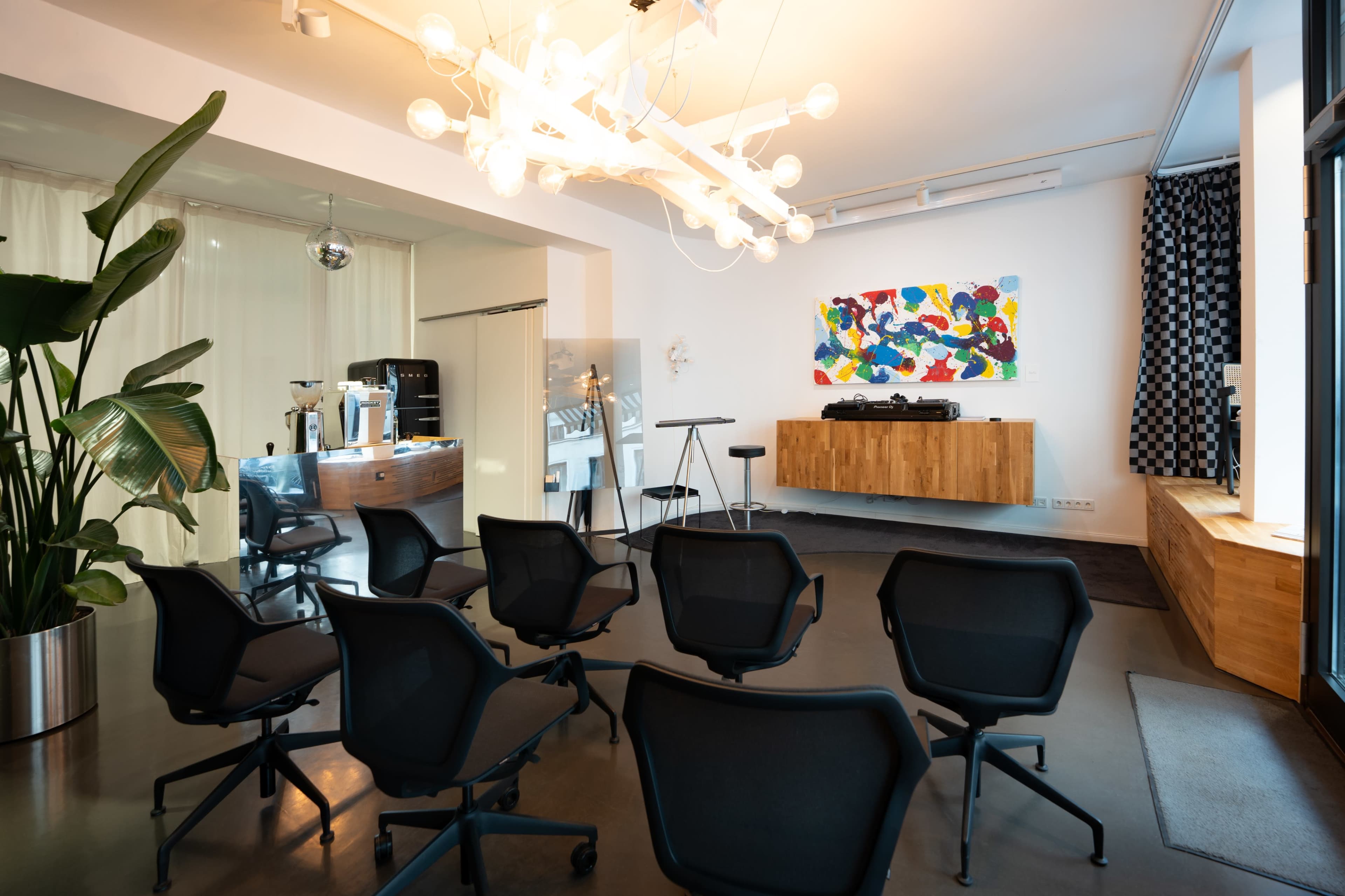 The image features a modern interior space with a seating arrangement of black chairs facing a wooden platform, vibrant abstract art on the wall, and a stylish light fixture overhead.