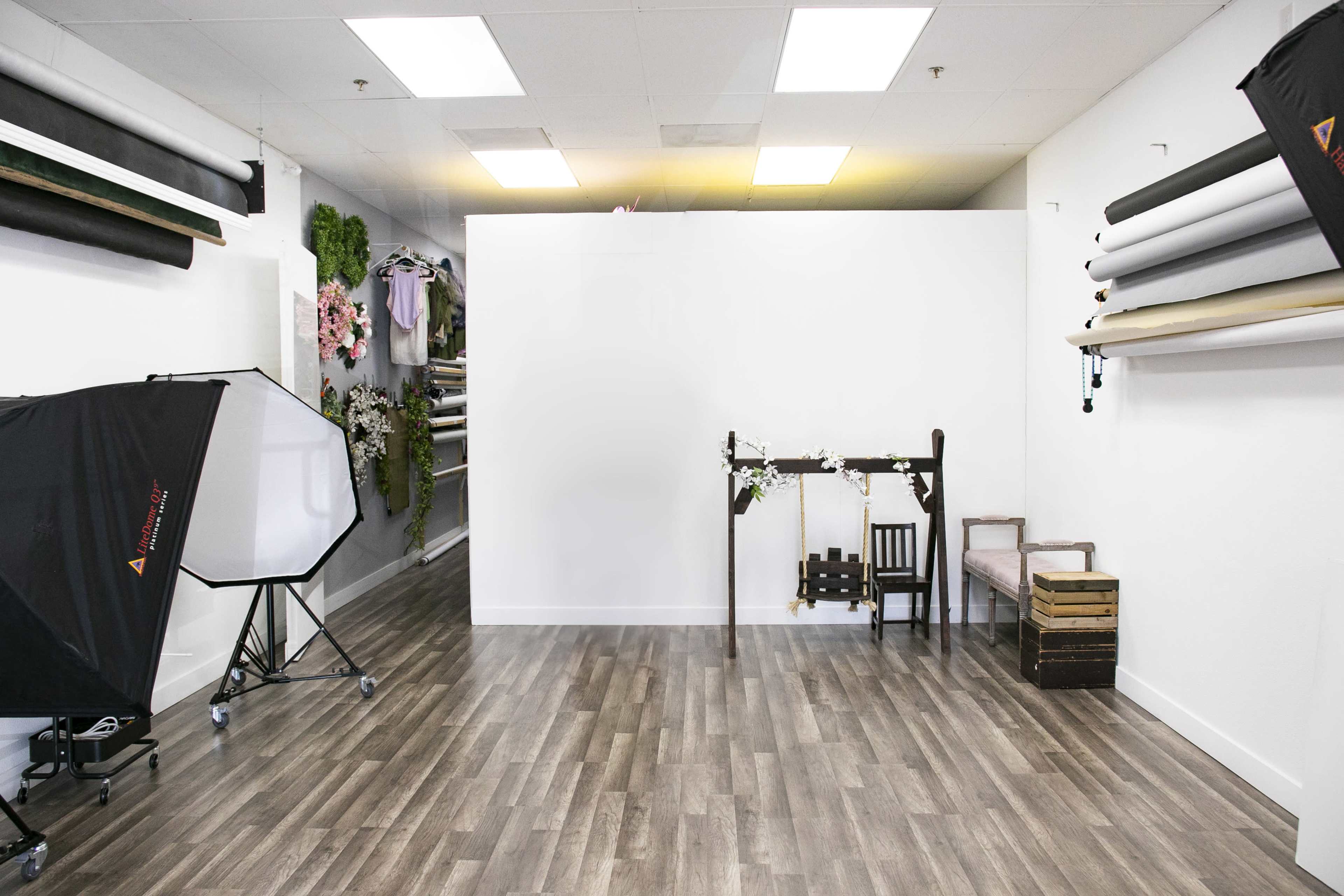 A photography studio featuring a blank white wall, a wooden table with a small chair, a vintage crate, and rolls of backdrop materials along the walls.