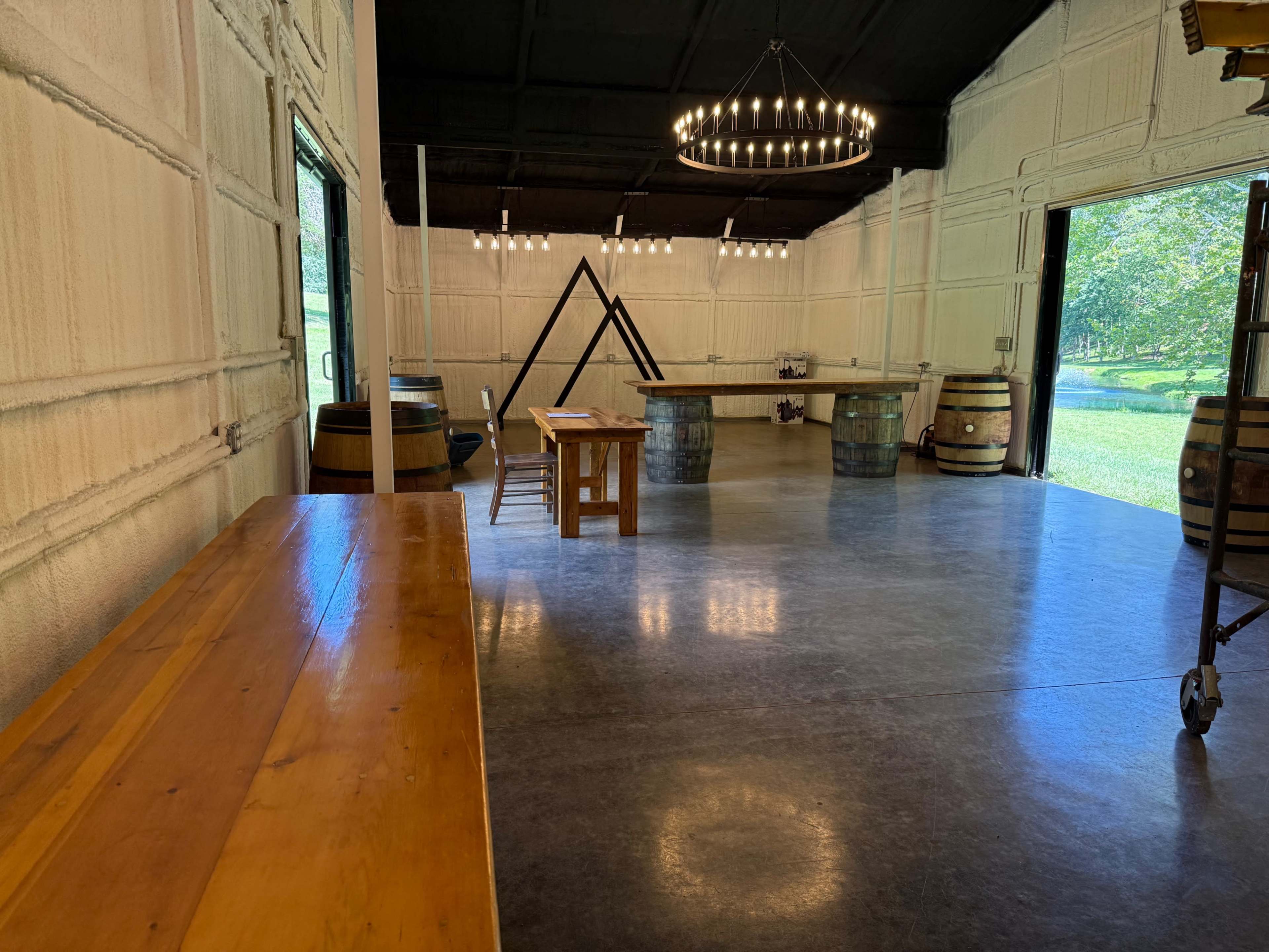 The image shows a spacious room with wooden furniture, large windows, and a high ceiling featuring a chandelier, along with wine barrels positioned around the space.