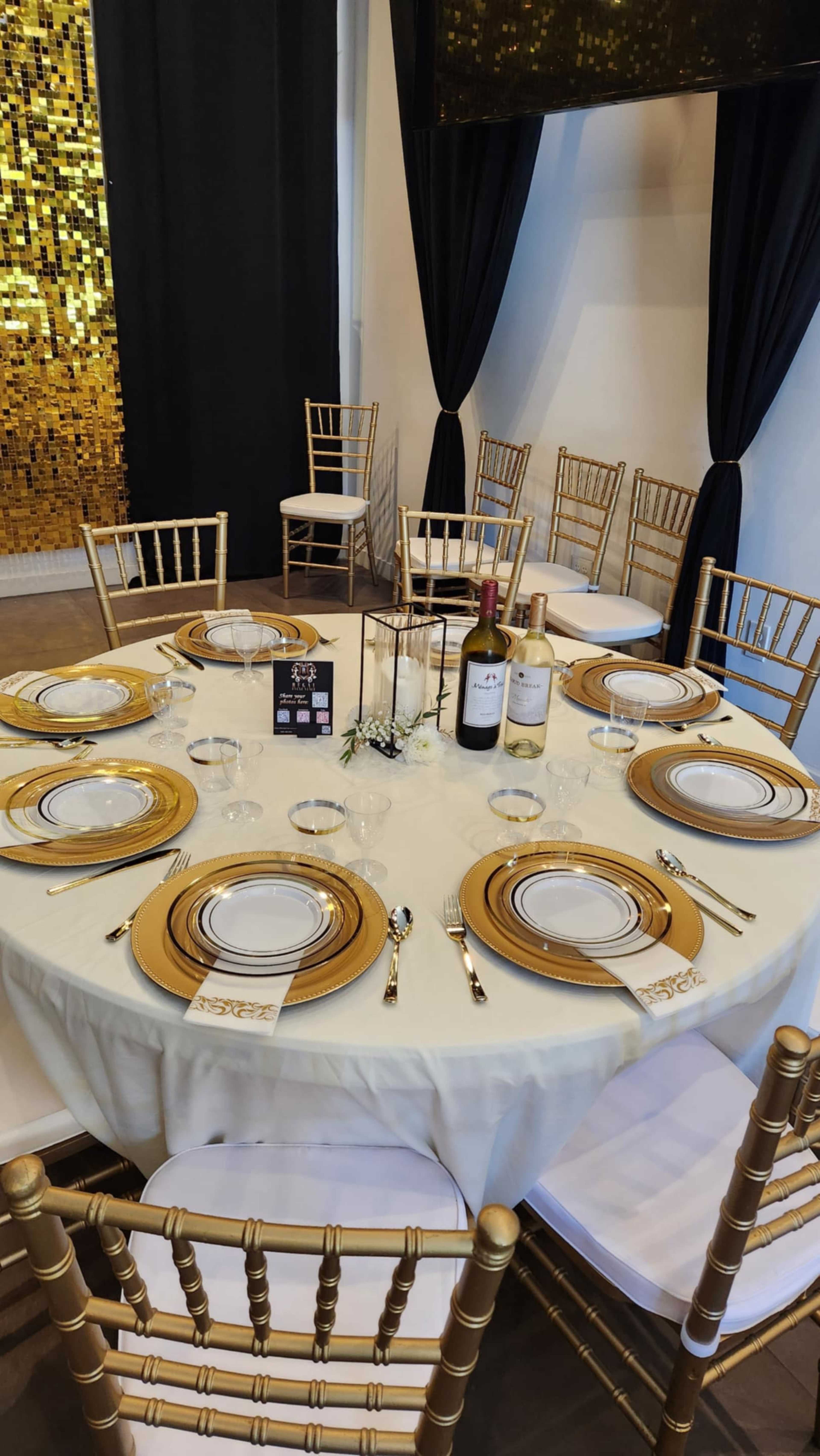 A round dining table is elegantly set with gold-rimmed plates, glassware, and wine bottles, surrounded by gold chiavari chairs.