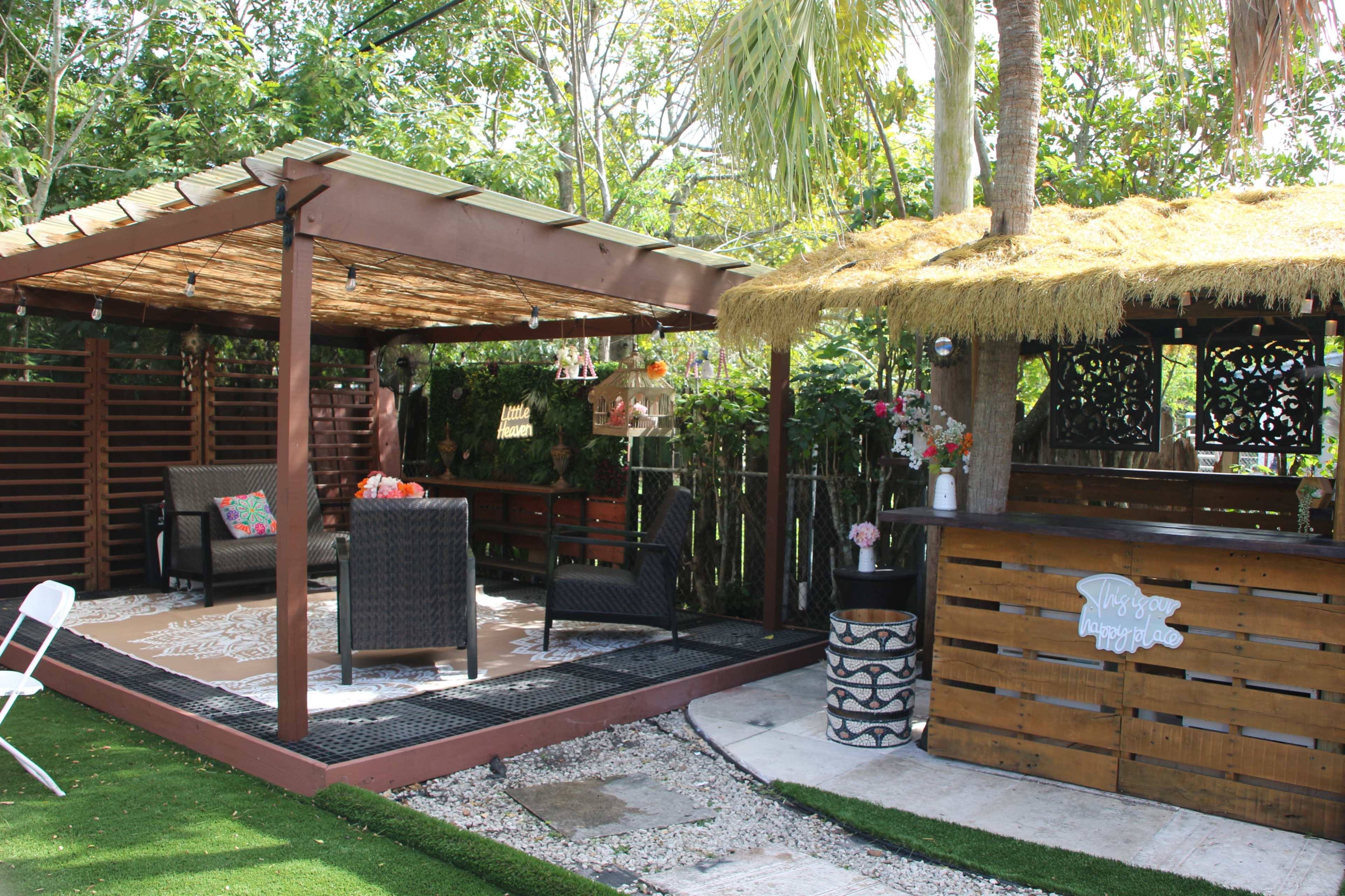 The image depicts a landscaped outdoor relaxation area featuring a covered wooden patio with seating, ornamental plants, and a small thatched-roof bar.