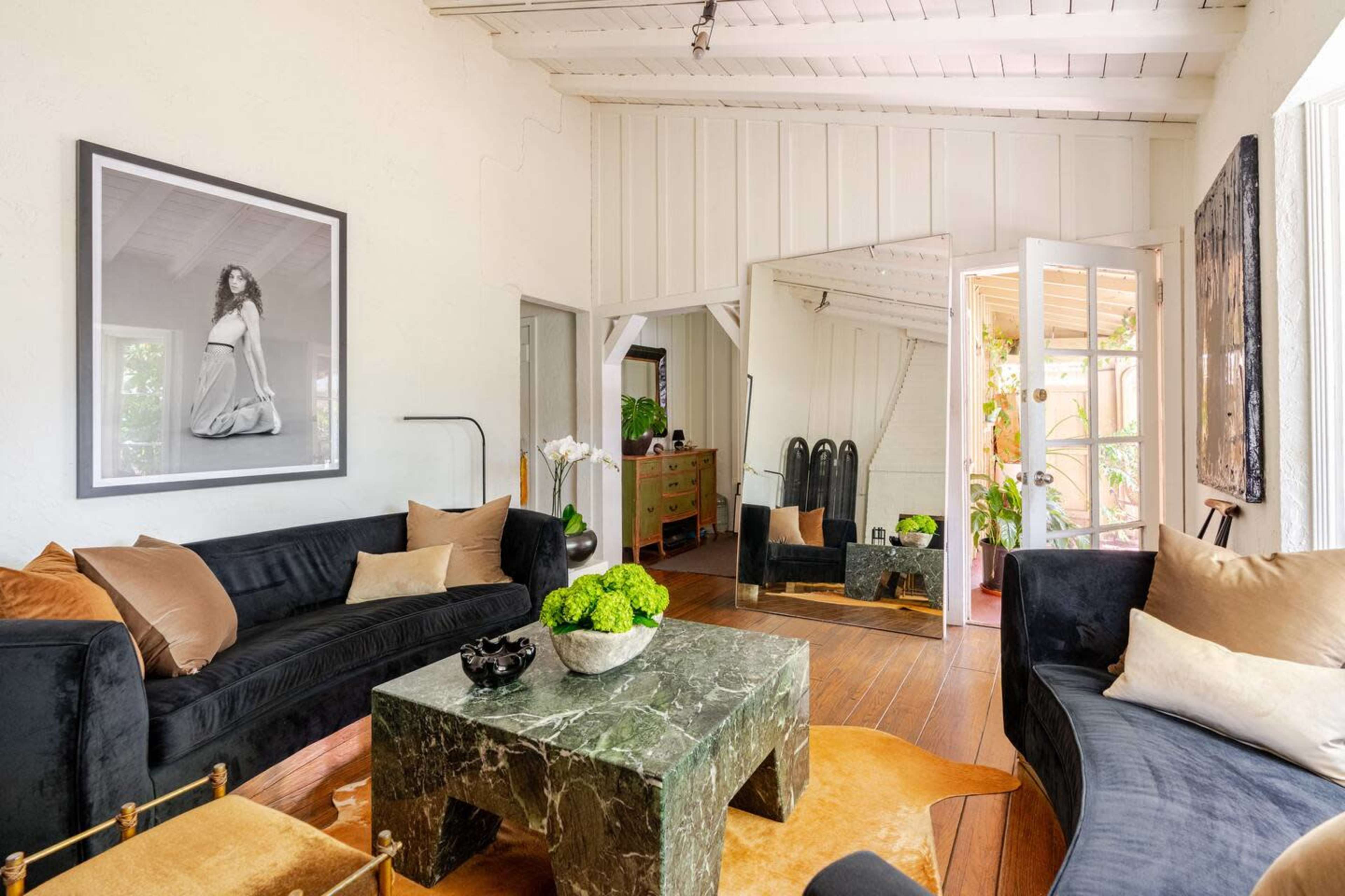The image shows a bright living room featuring a dark velvet sofa, a marble coffee table, a large framed photograph, and an open door leading to a patio with plants.