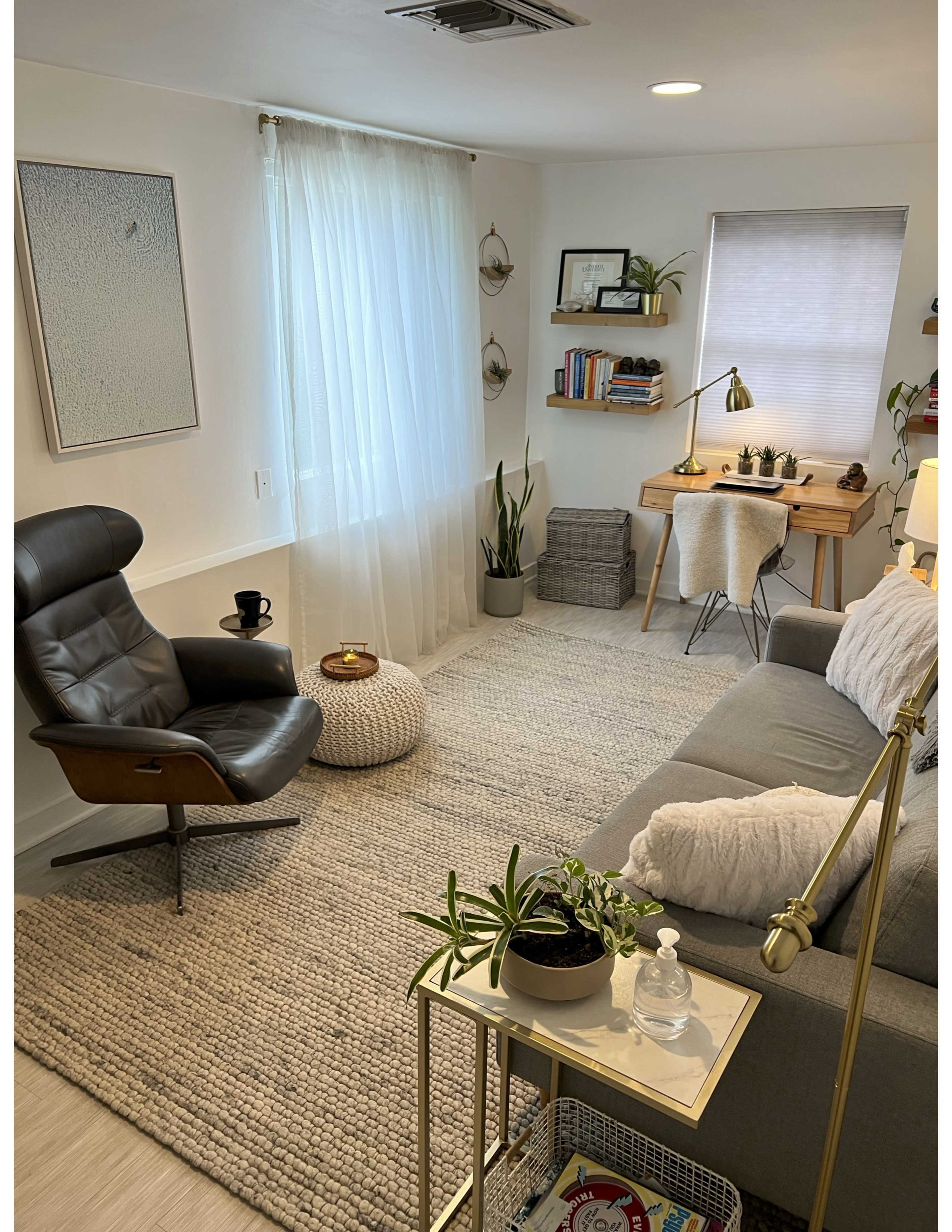 The image shows a cozy living room with a gray sofa, a black armchair, a small coffee table, and a wooden desk, all arranged around a soft rug under natural light from a window.