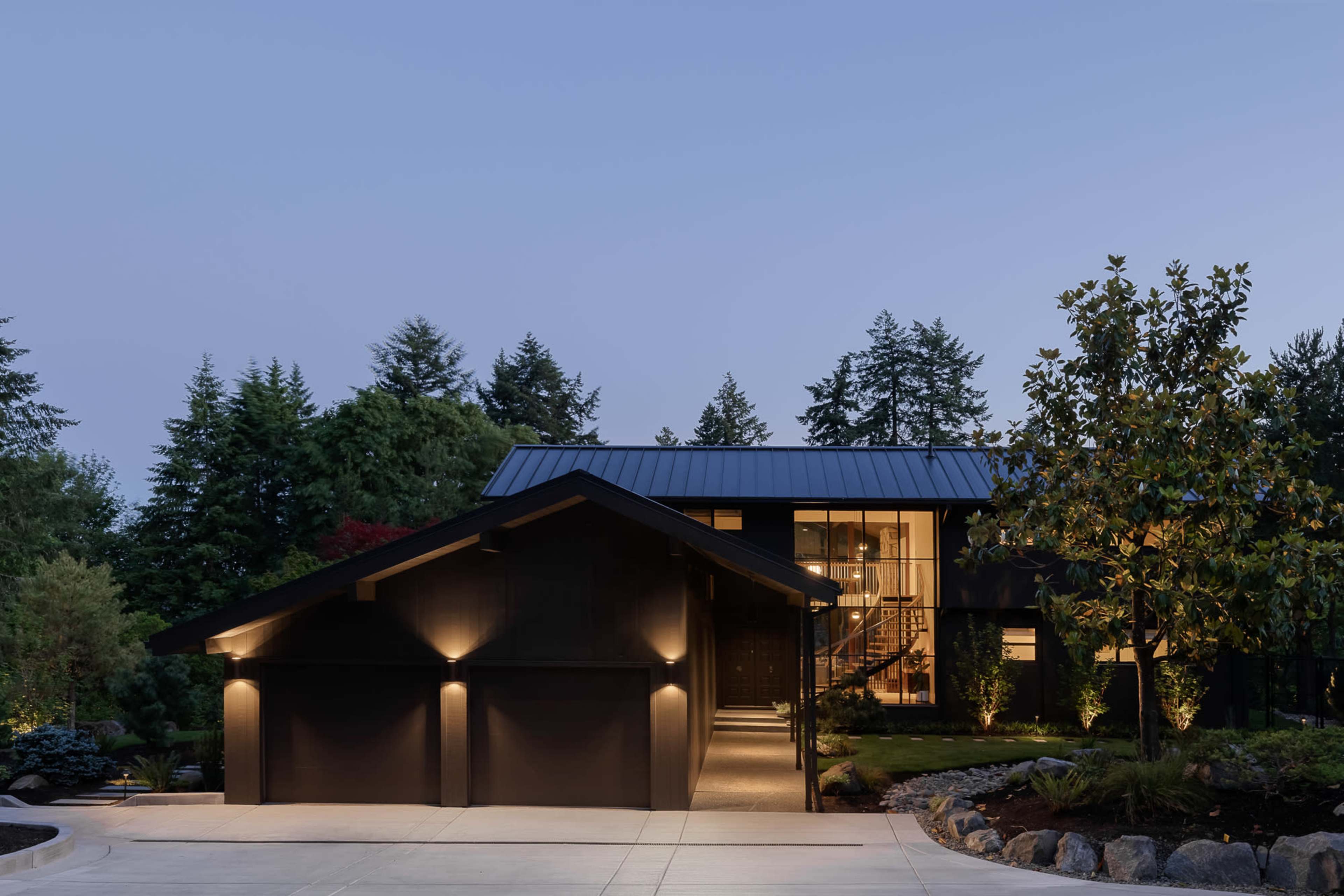 A contemporary house with a dark exterior and large windows is illuminated at dusk, surrounded by trees and a landscaped driveway.