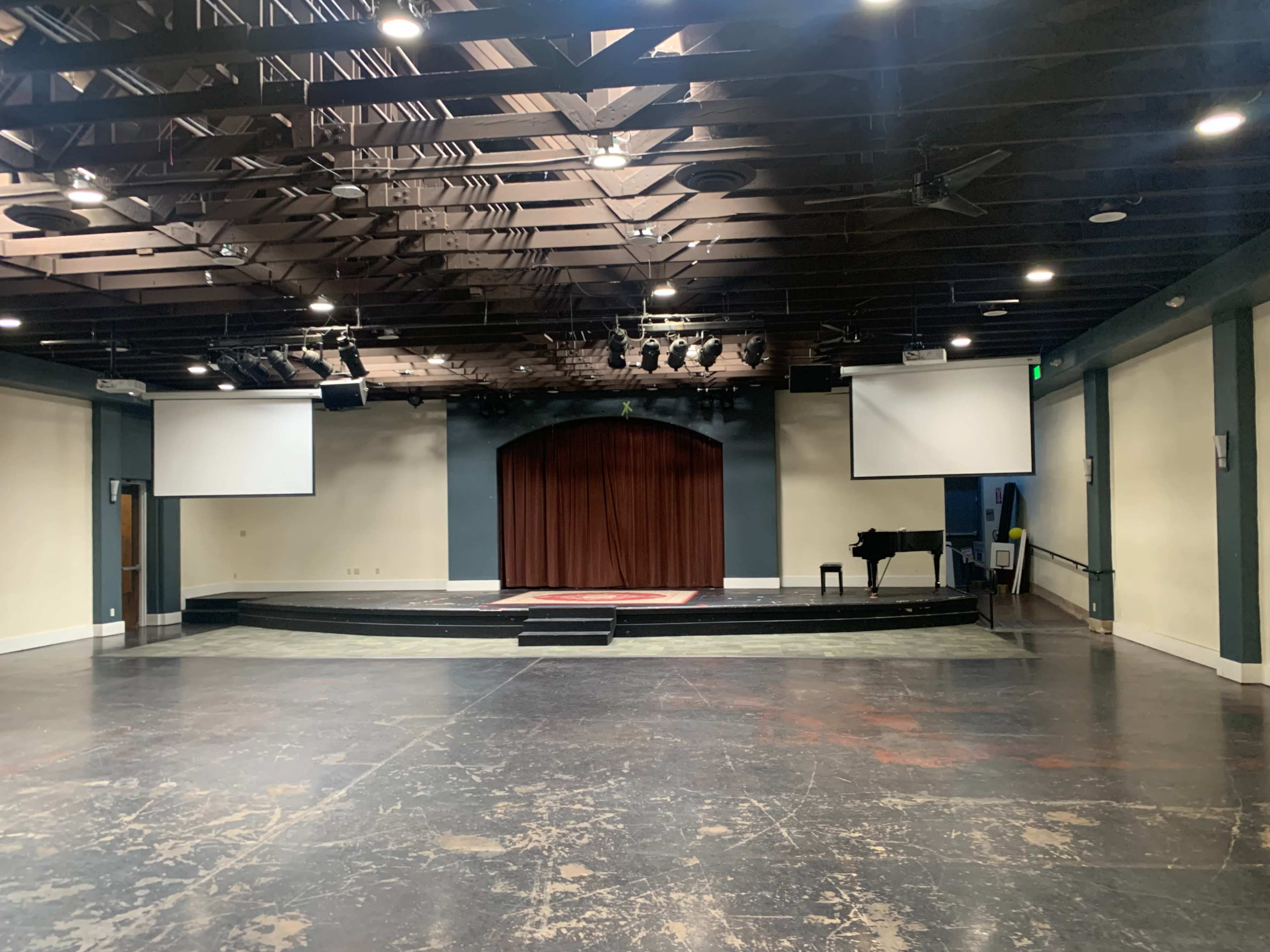 The image shows a stage area in a performance space featuring a dark curtain, two projection screens, and a piano, all within a room with a wooden ceiling.