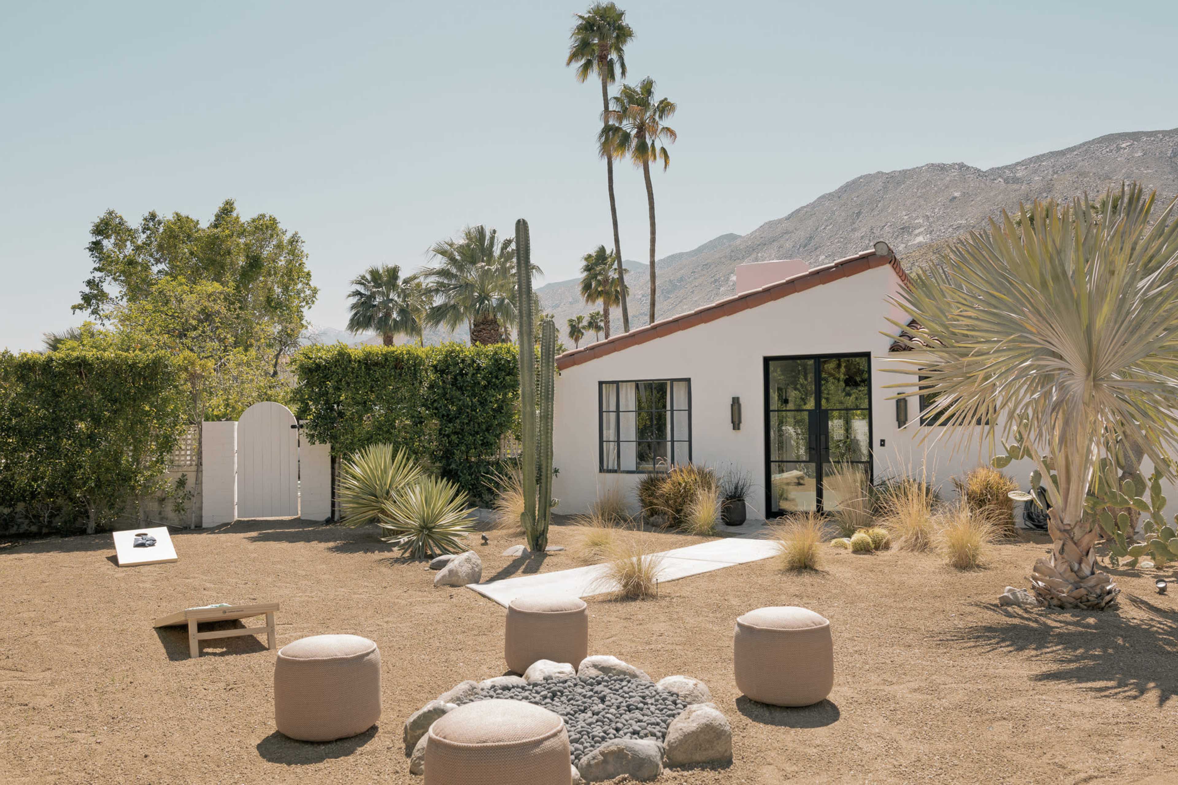A desert landscape with a small, white house surrounded by palm trees, desert plants, and a circular seating arrangement, set against a backdrop of mountains.