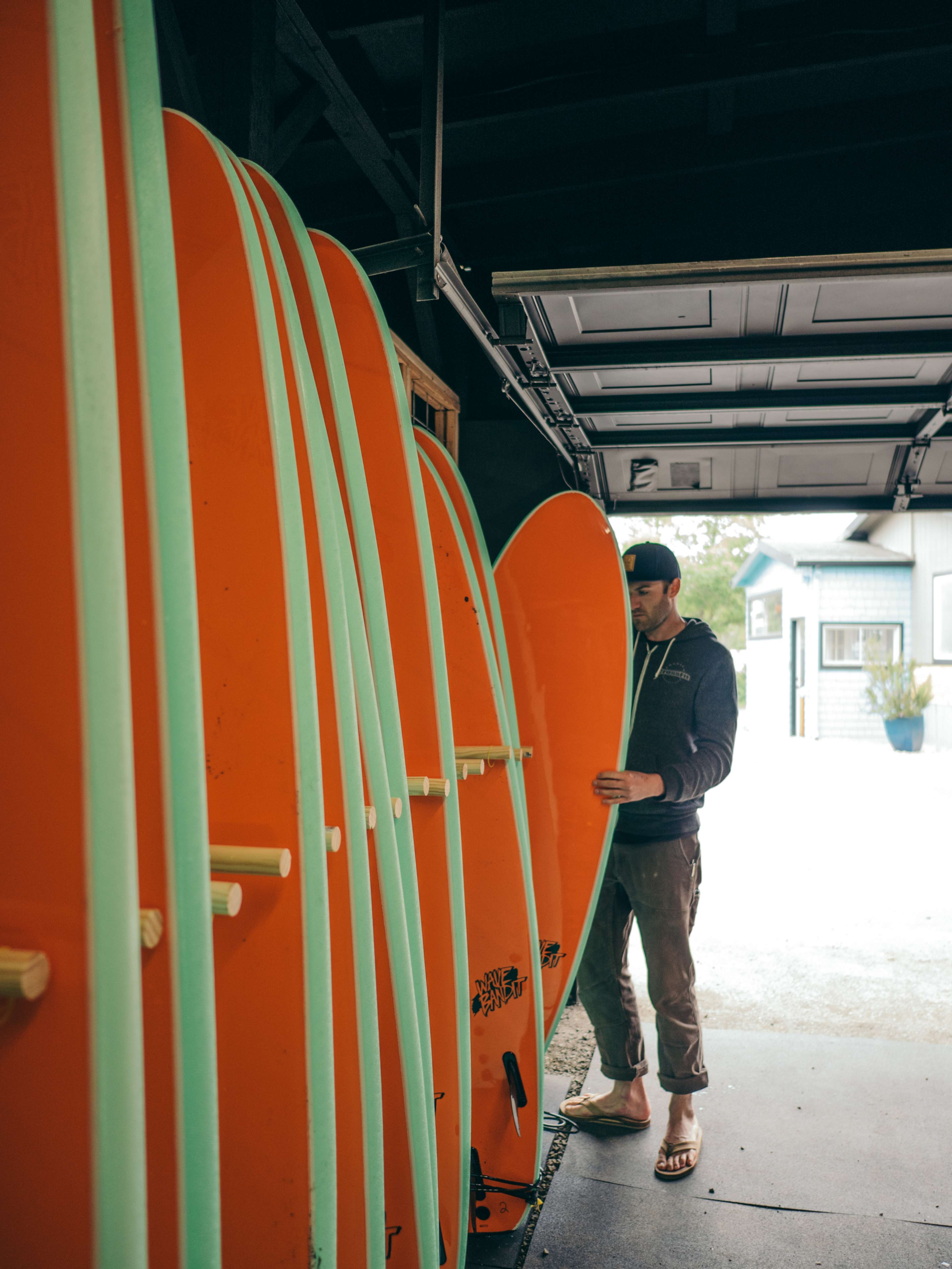 A man stands in a garage, arranging vibrant orange surfboards with teal accents.