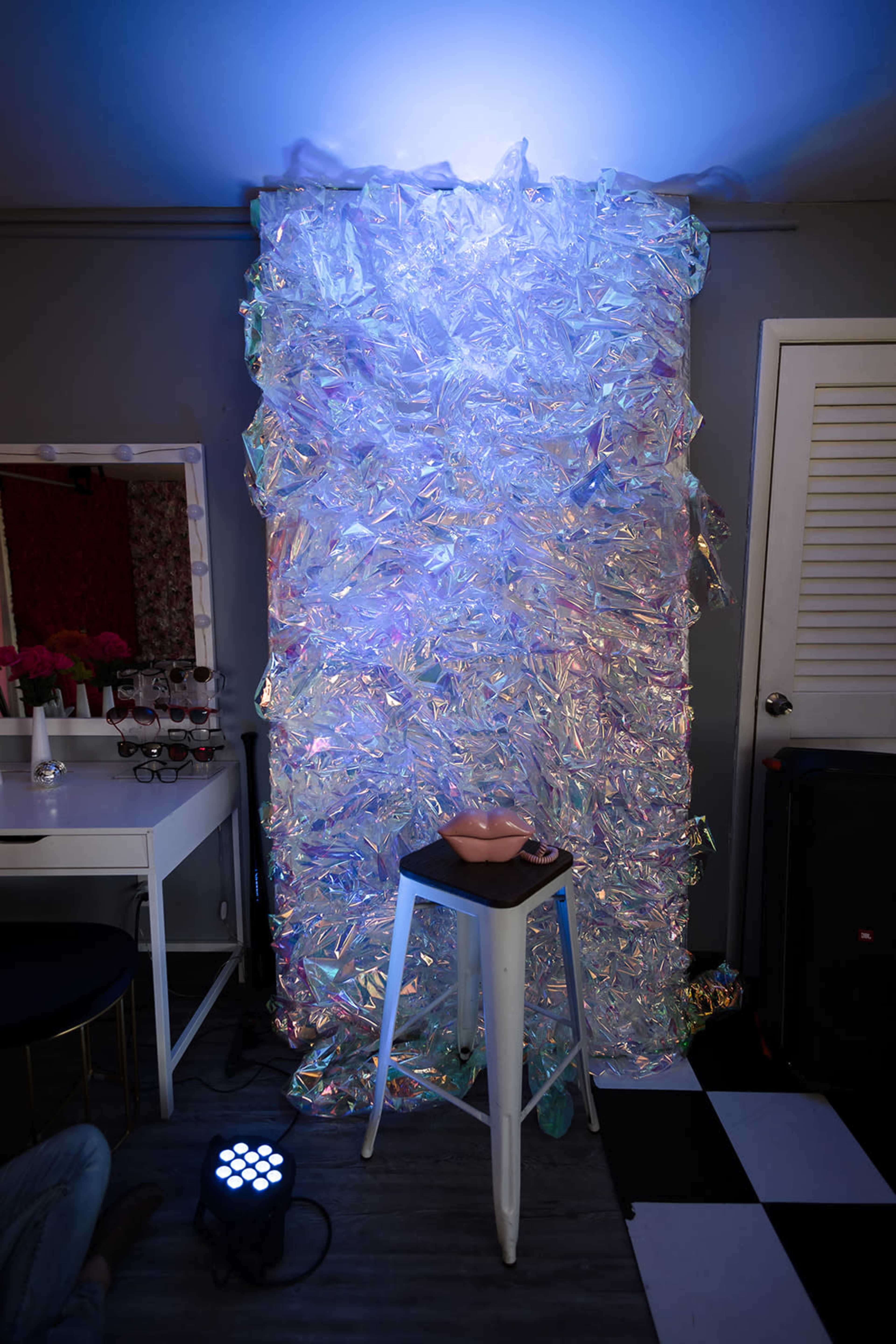 A translucent, iridescent plastic backdrop is illuminated by a blue light, standing behind a small white table topped with a bowl, in a room with black and white checkered flooring.