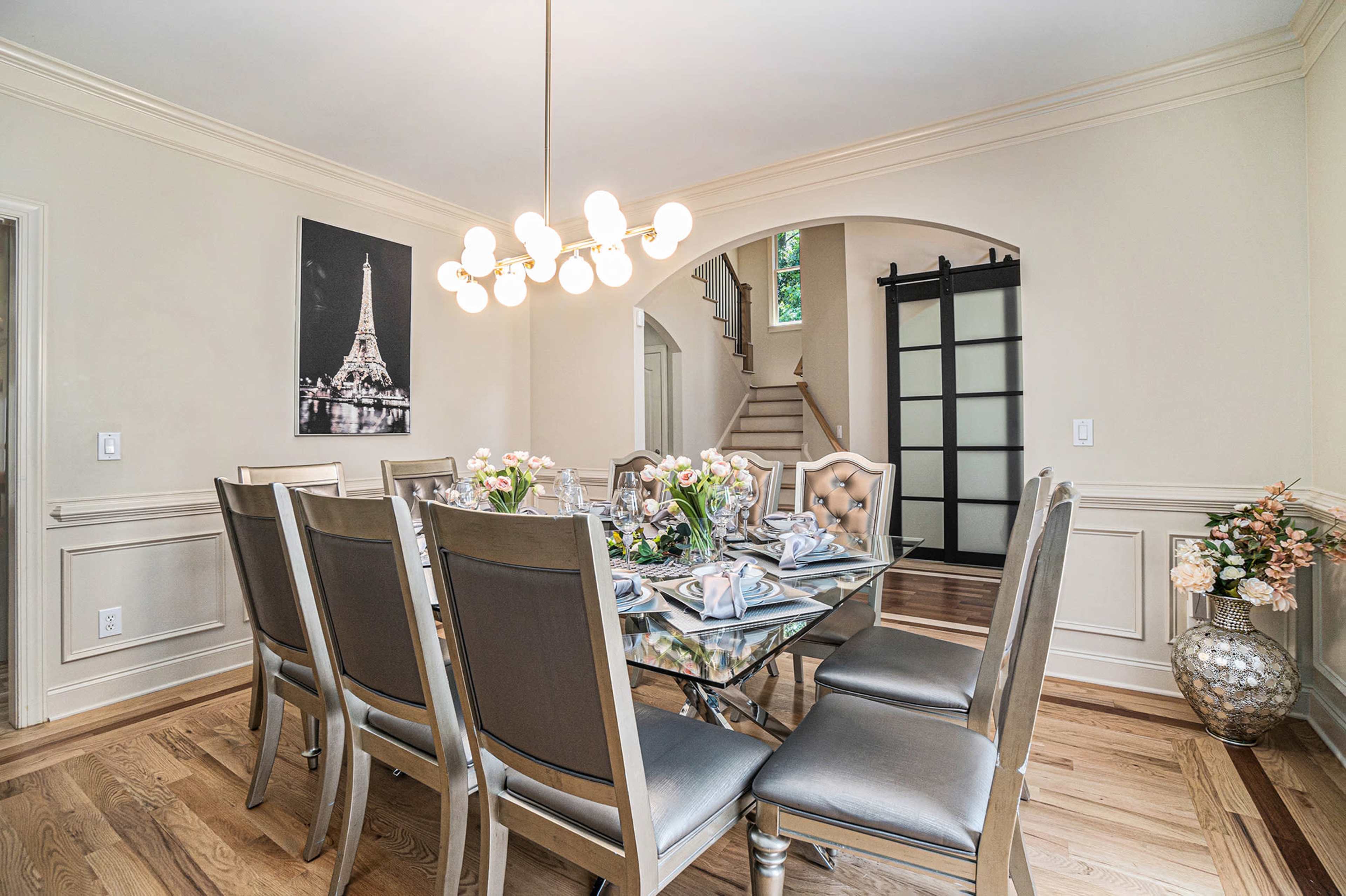A modern dining room features a glass table set with floral arrangements, surrounded by elegantly designed chairs, with a large archway leading to a staircase.