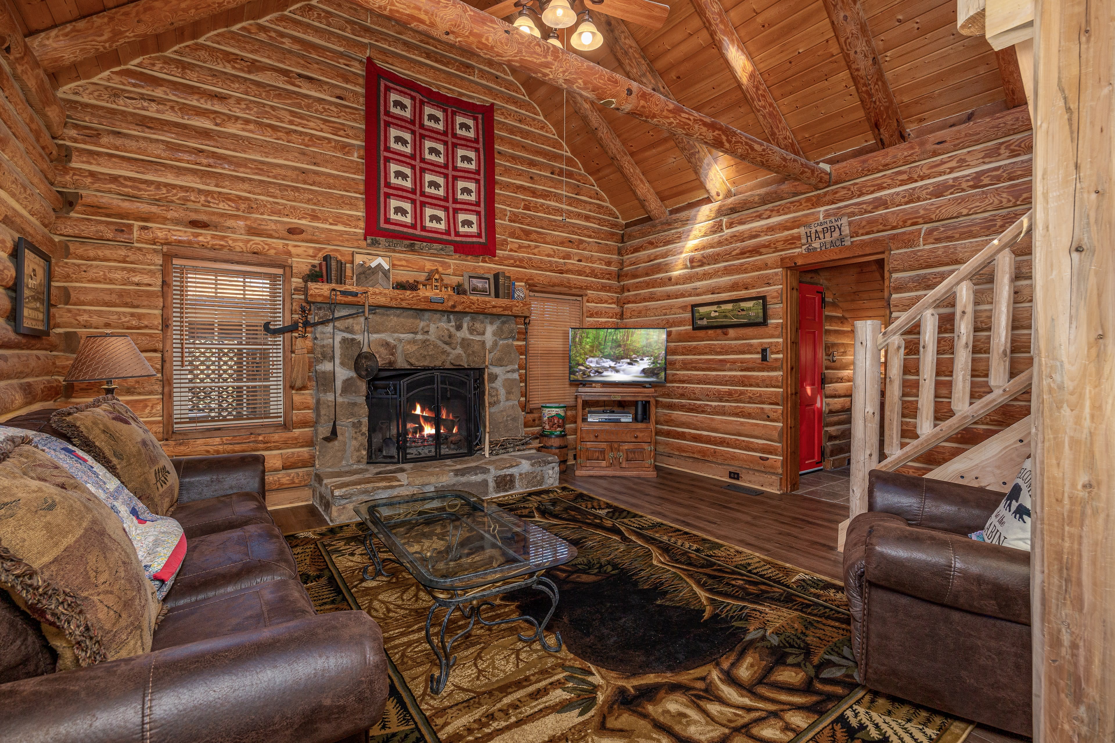 The image shows a rustic log cabin living room with a stone fireplace, wooden beams, and a staircase leading to a second level.